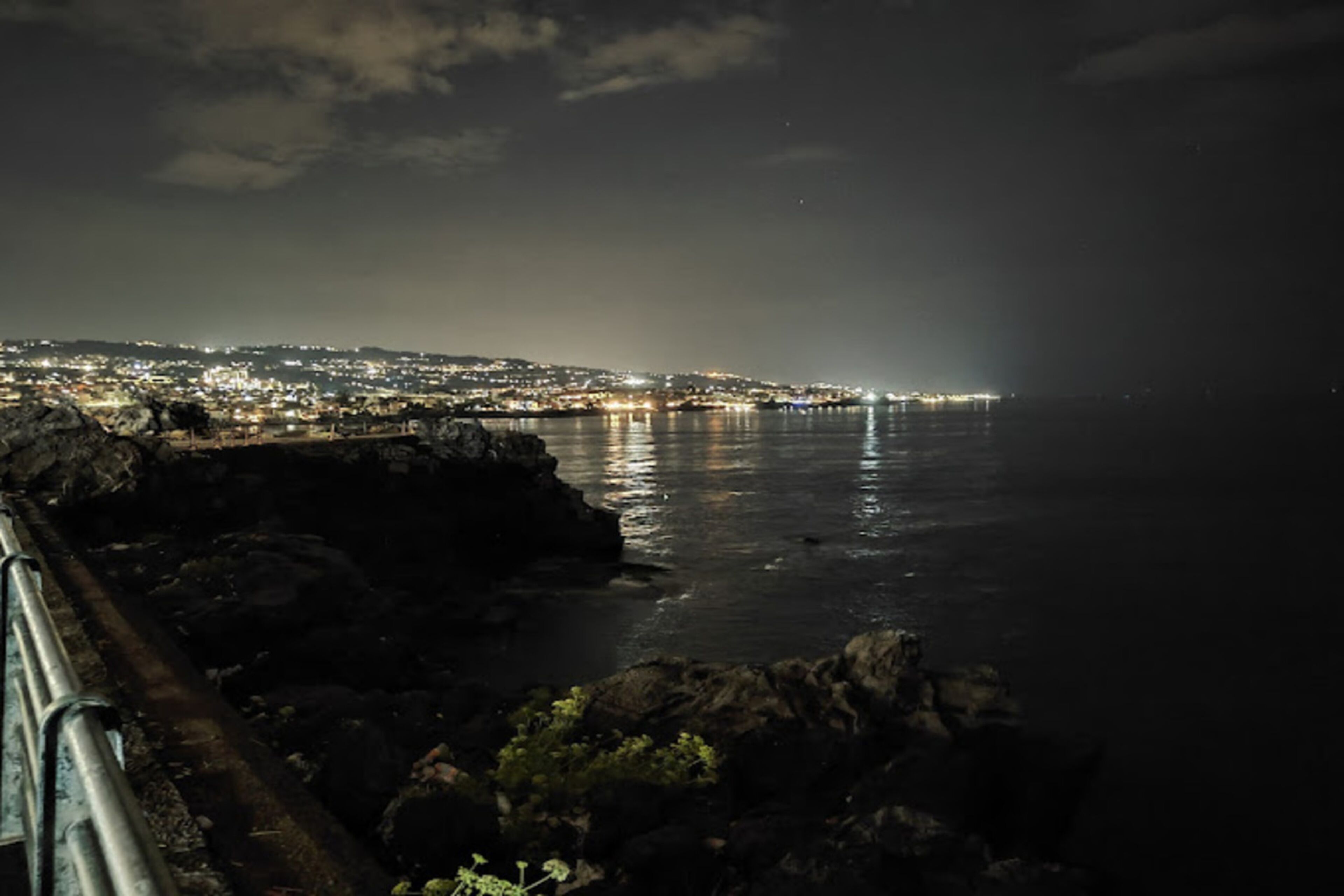 Panoramica costiera notturna con un litorale roccioso e un molo che si protende sull'acqua.