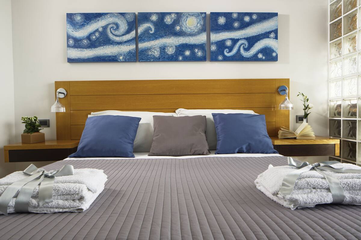A neatly made bed with a grey quilt, two blue pillows, and a brown pillow. There are two bedside tables with lamps and a small plant. The room has a wooden headboard and three blue paintings on the wall.