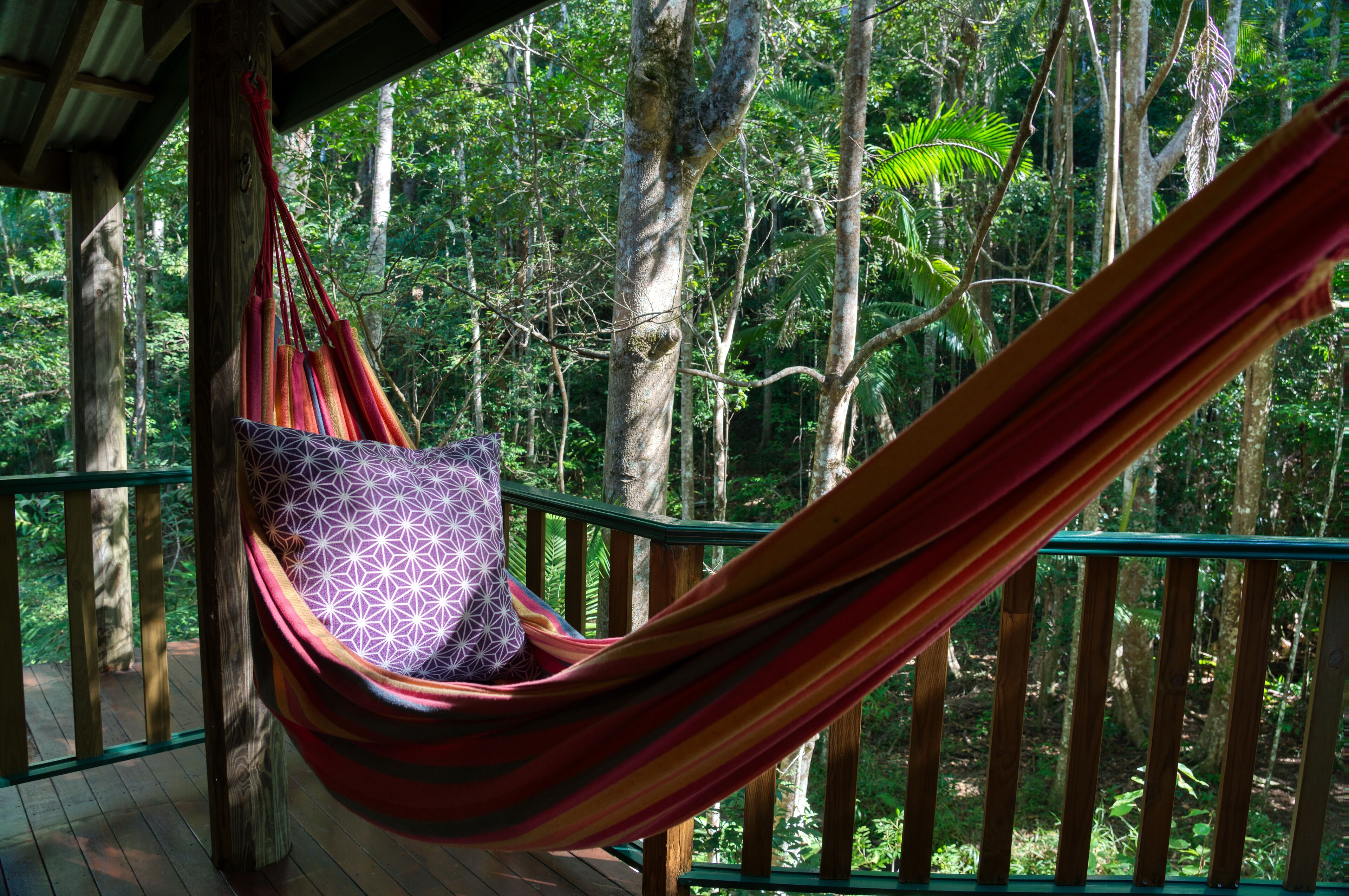 A hammock with a patterned cushion in a forested area with a wooden deck and railing.
