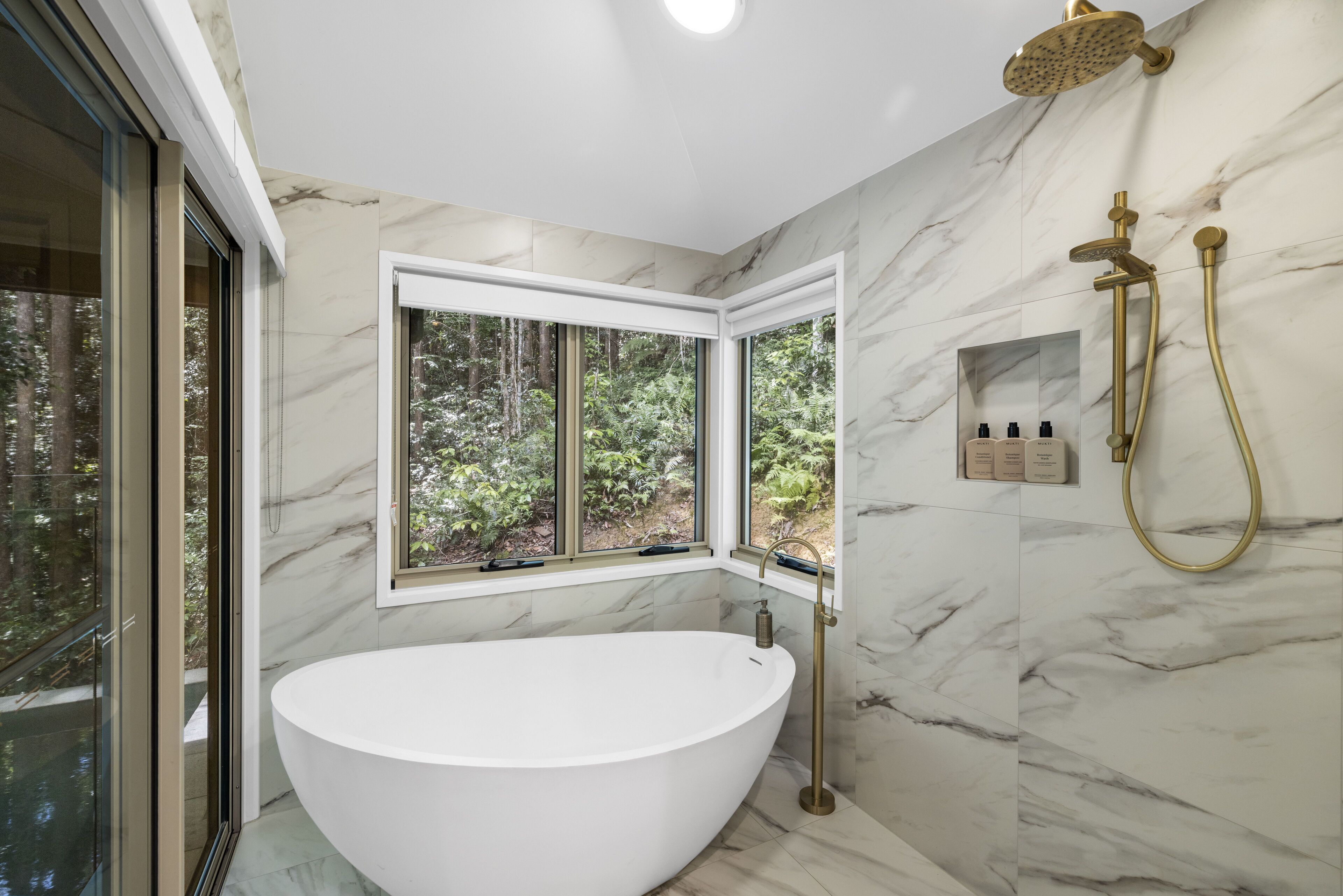 A modern bathroom with a freestanding bathtub, a showerhead, and a window with a view of greenery.