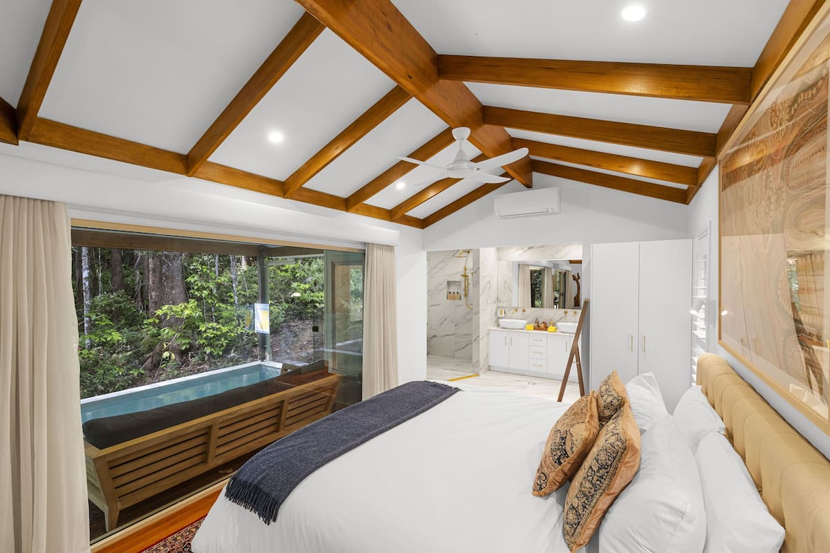 A bedroom with a large bed, wooden beams, a ceiling fan, a view of a forest through a glass door, and a bathroom with a marble sink.