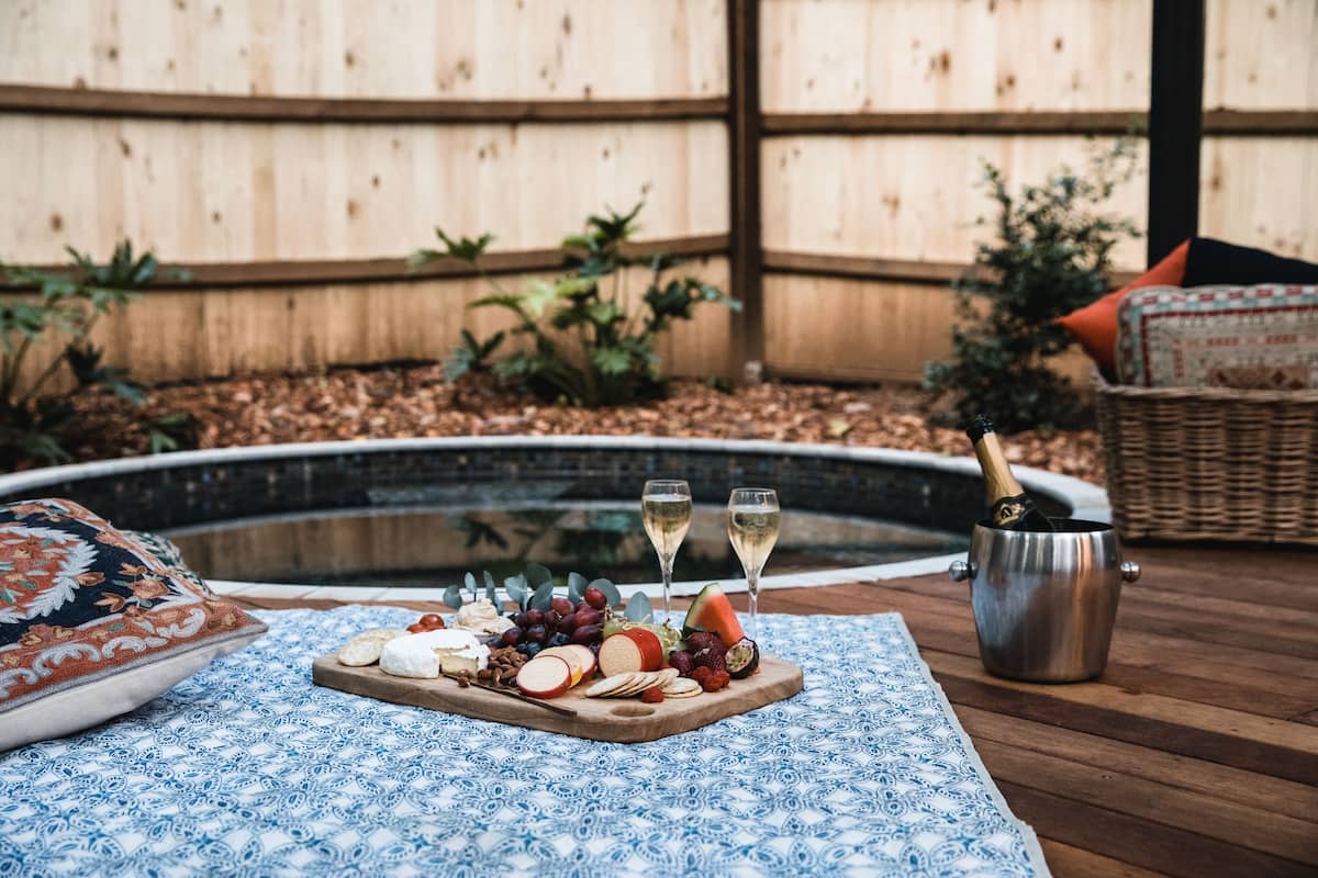 A wooden deck with a picnic setup including a wooden board with cheese, grapes, and a watermelon, two champagne glasses, and a metal bucket with a bottle.