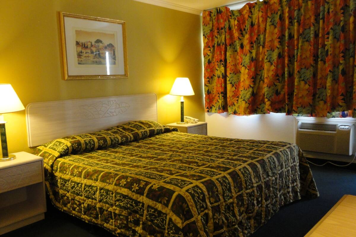 A hotel room with a bed, two bedside lamps, a framed picture on the wall, and a window with floral curtains.