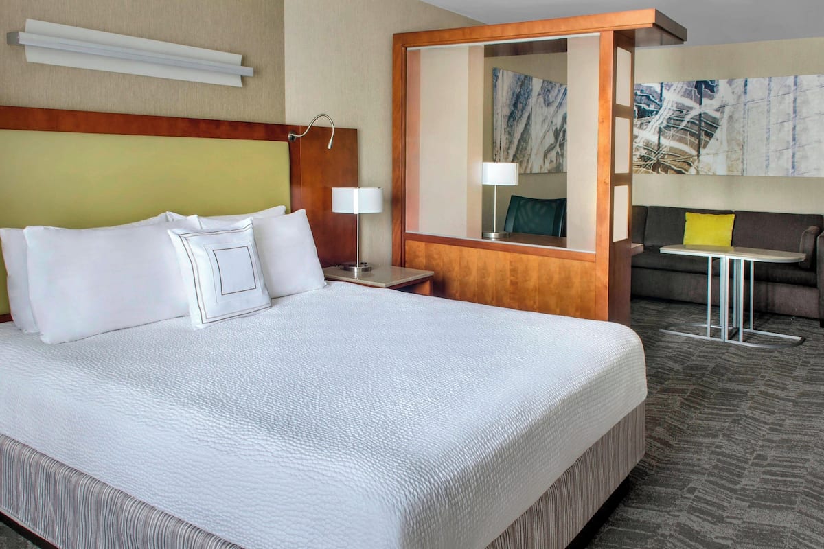 Habitación de hotel con una cama grande, una mesita de noche con lámpara, una pequeña mesa con silla y un espejo amplio.