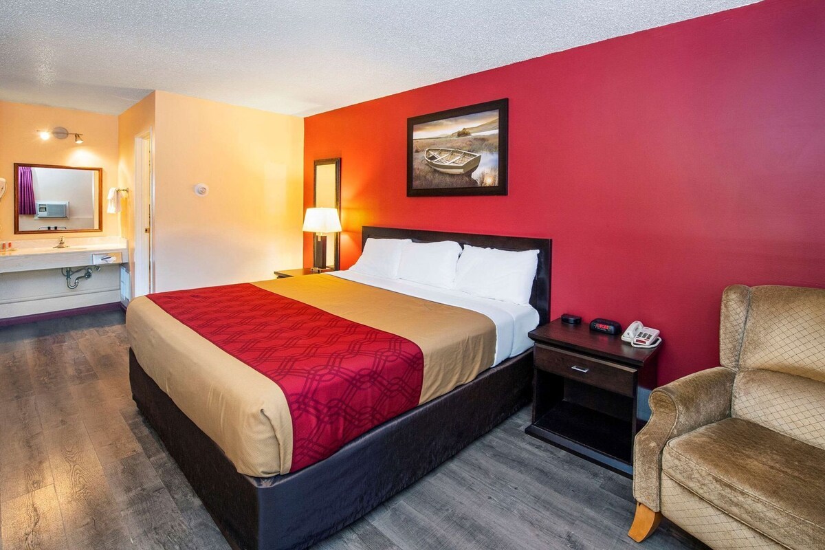 A hotel room with a bed, a nightstand, a chair, a bathroom with a mirror, and a painting on the wall.