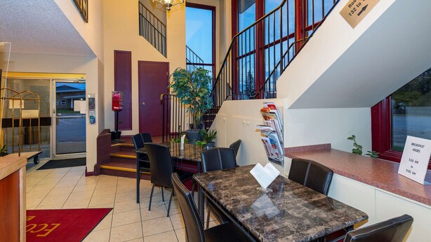 A hotel lobby with a reception desk, a dining area with a marble table and chairs, a staircase, and a sign indicating breakfast hours. - Econo Lodge Edson - 2nd Ave (Edson)