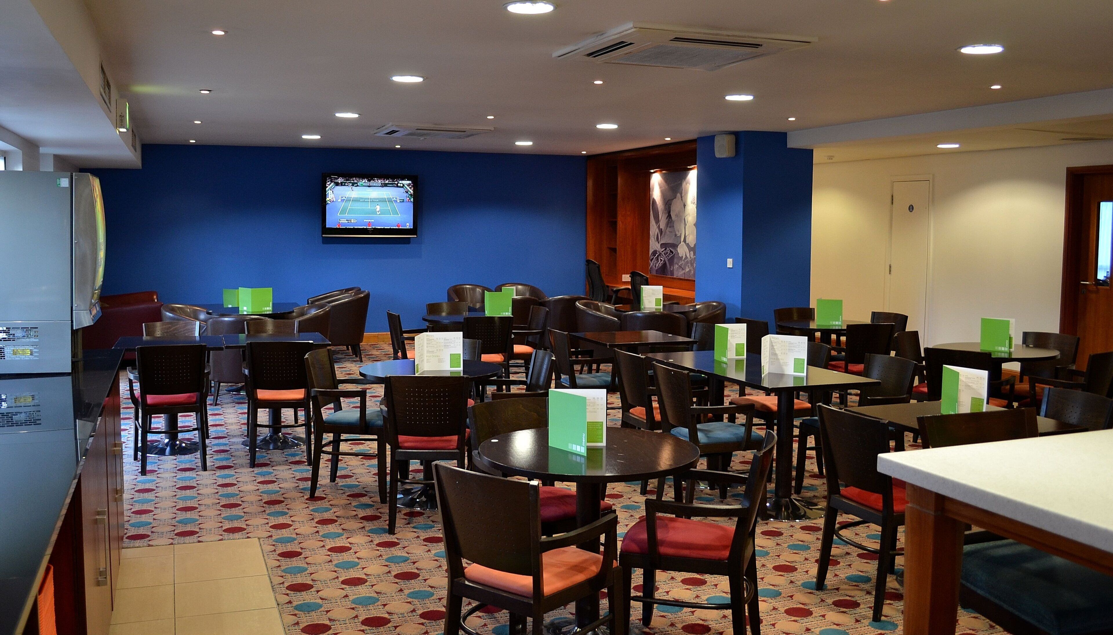 Comedor de hotel con mesas y sillas, una pantalla de televisión y una alfombra estampada.