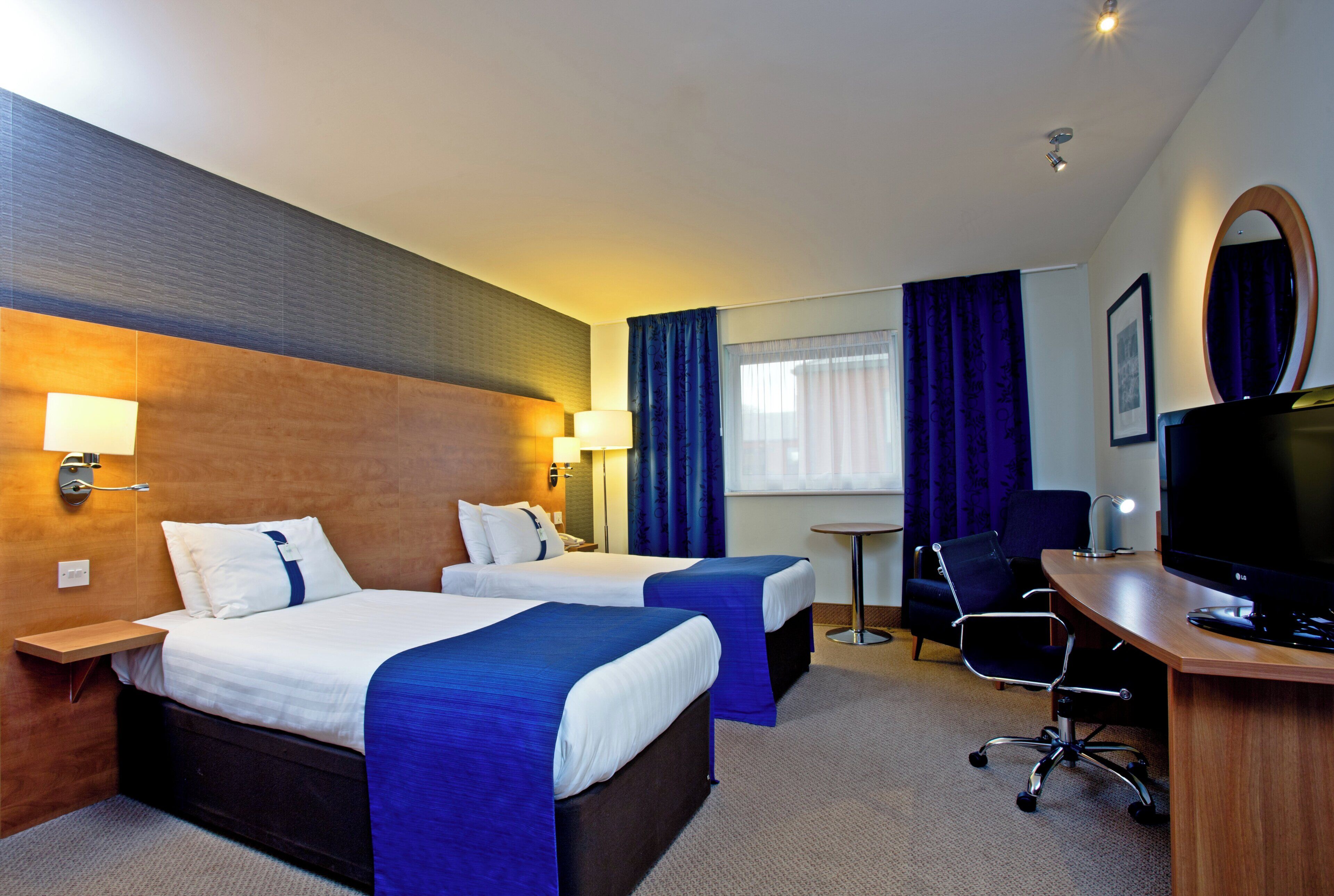 A hotel room with two beds, a desk, a chair, a TV, and a small table.