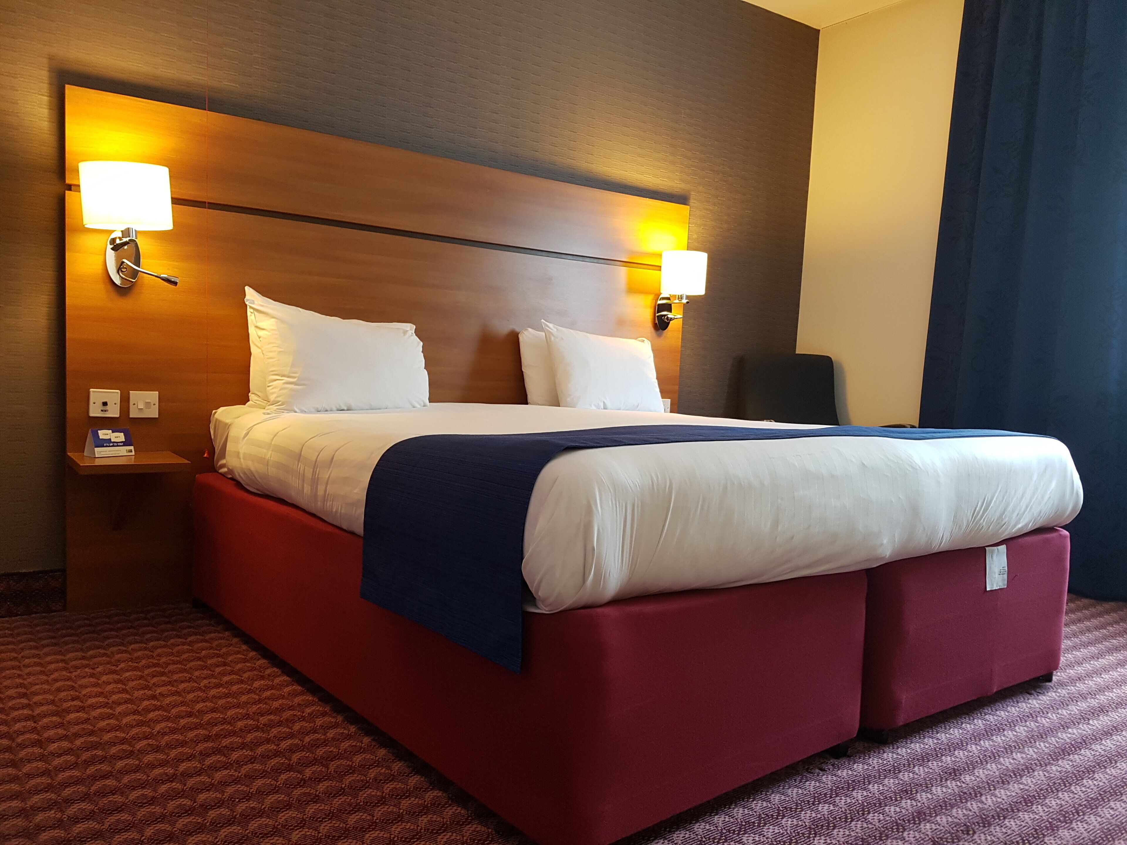 A hotel room with a large bed, two bedside lamps, a nightstand, and a red carpet.