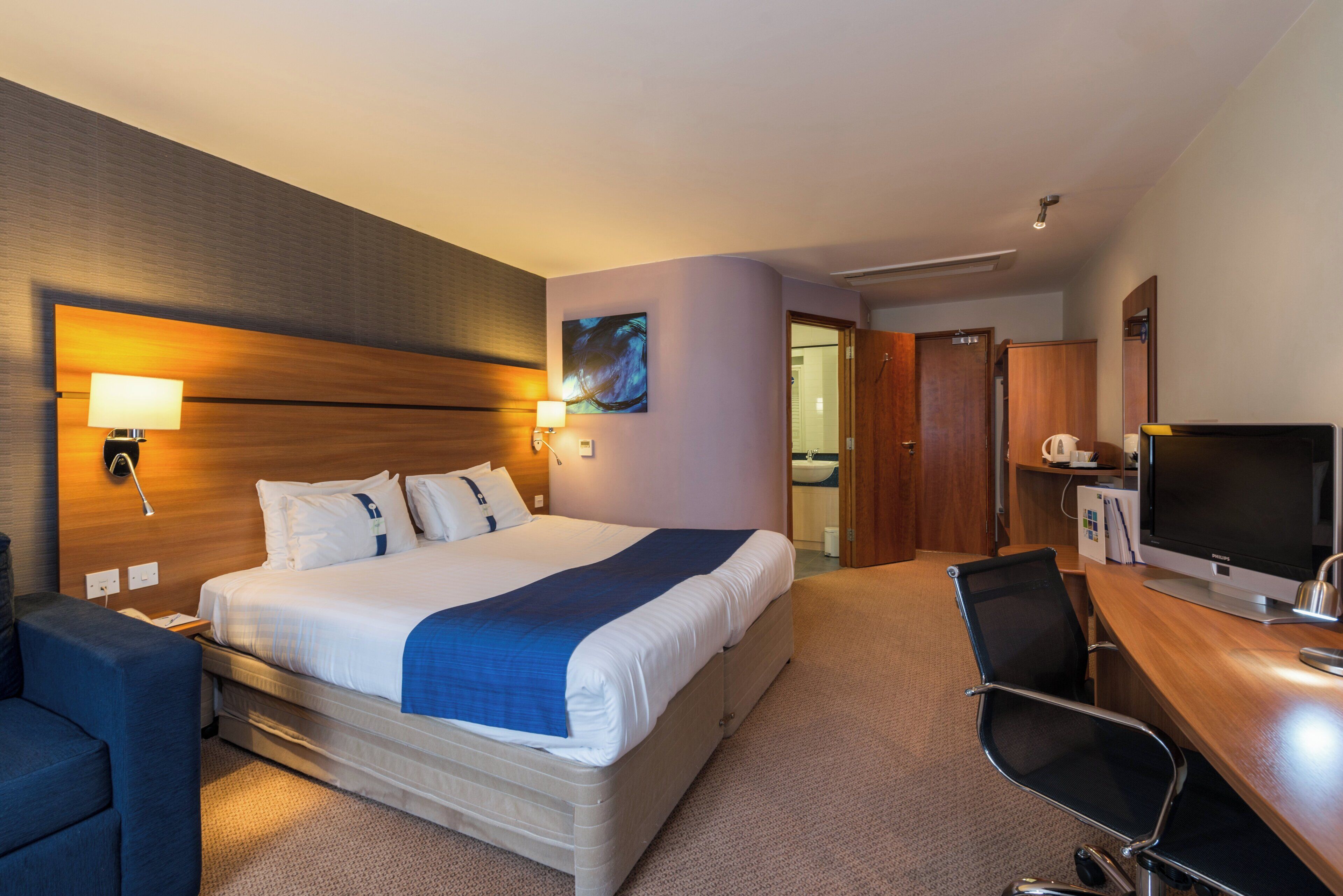 Una habitación de hotel con una cama grande, un escritorio con computadora, una silla, un televisor y un baño visible a través de una puerta abierta.
