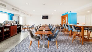 A modern dining area with multiple tables and chairs, a kitchen area with a refrigerator and coffee machine, and a blue accent wall.