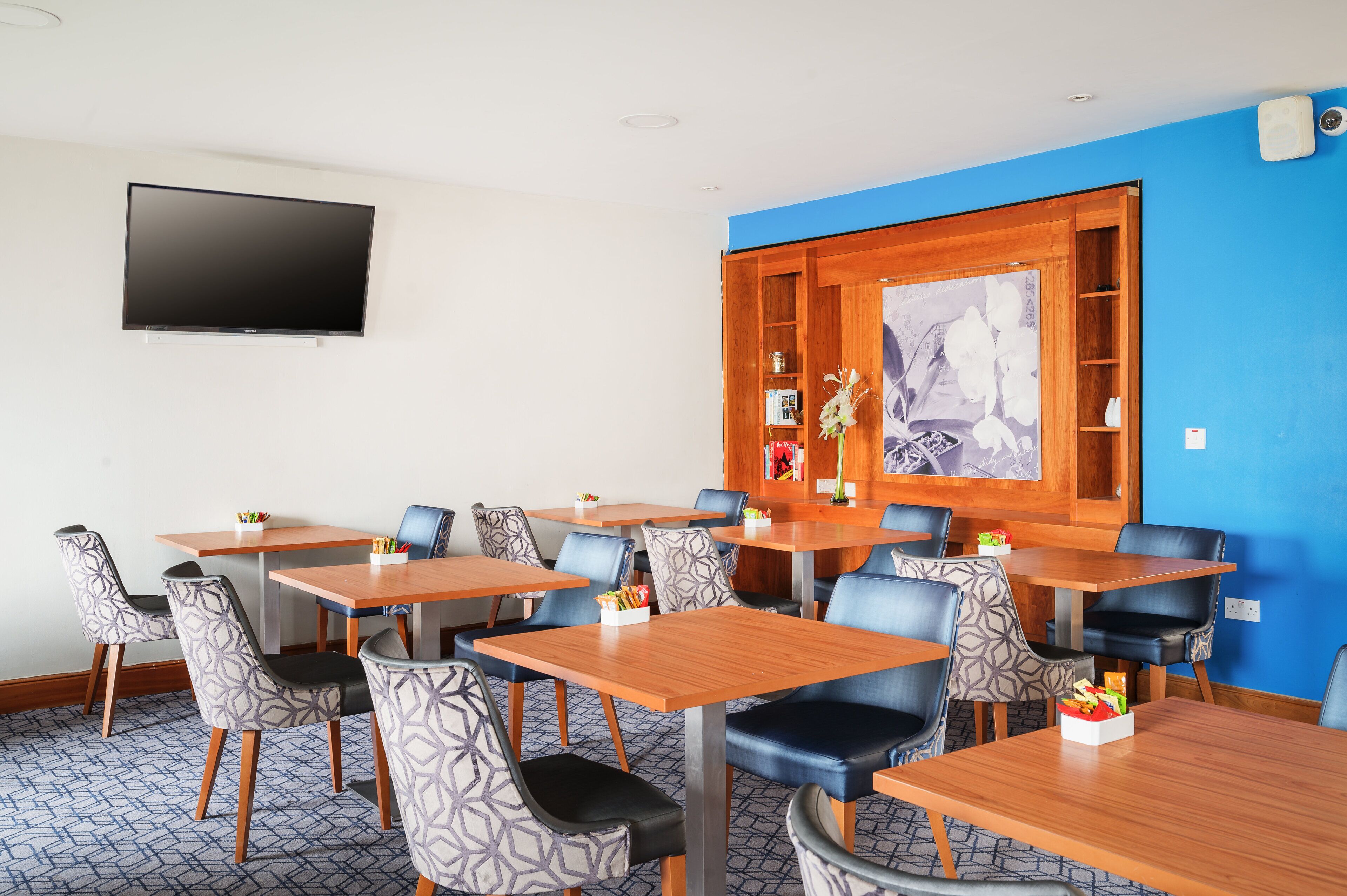 A hotel room with a flat-screen TV mounted on the wall, a wooden cabinet with a floral painting, and a blue accent wall.