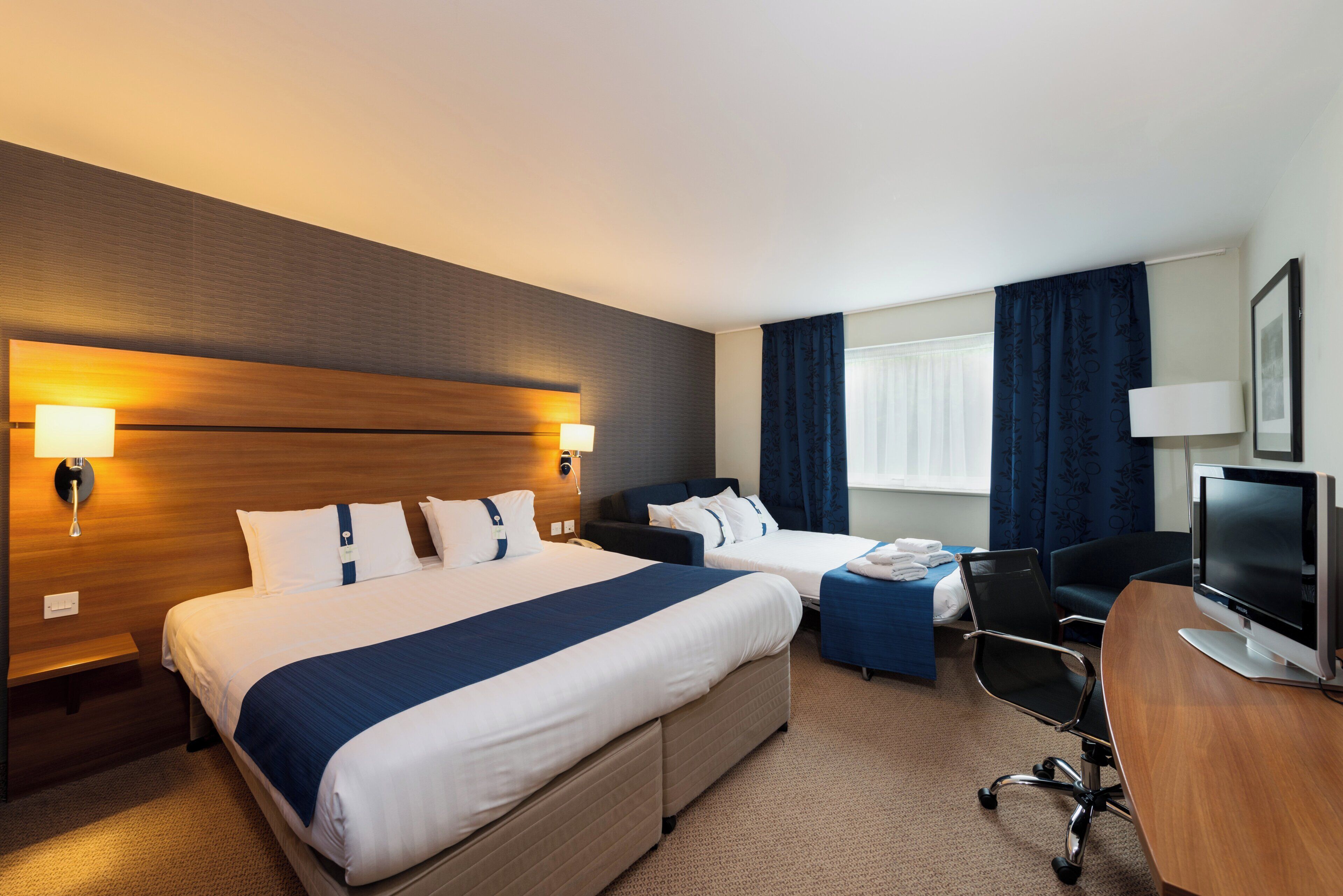 A hotel room with two beds, a desk, a chair, a television, and a window with curtains.