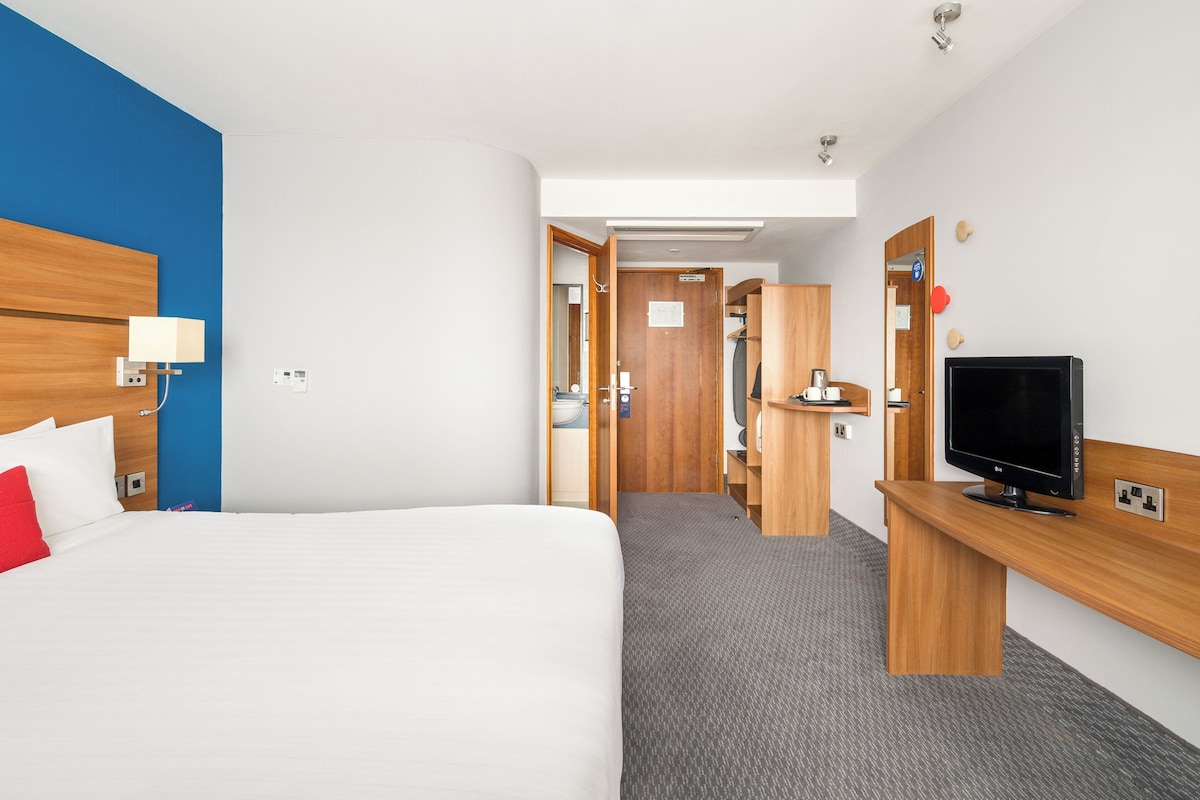 A hotel room with a large bed, a desk with a flat-screen TV, a wooden headboard, and a blue accent wall.