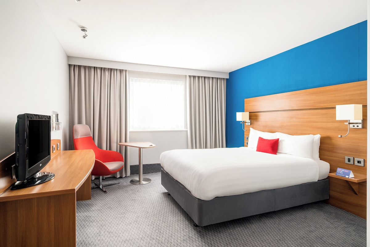 A hotel room with a bed, a desk with a TV, a small table, a red chair, and a blue accent wall.