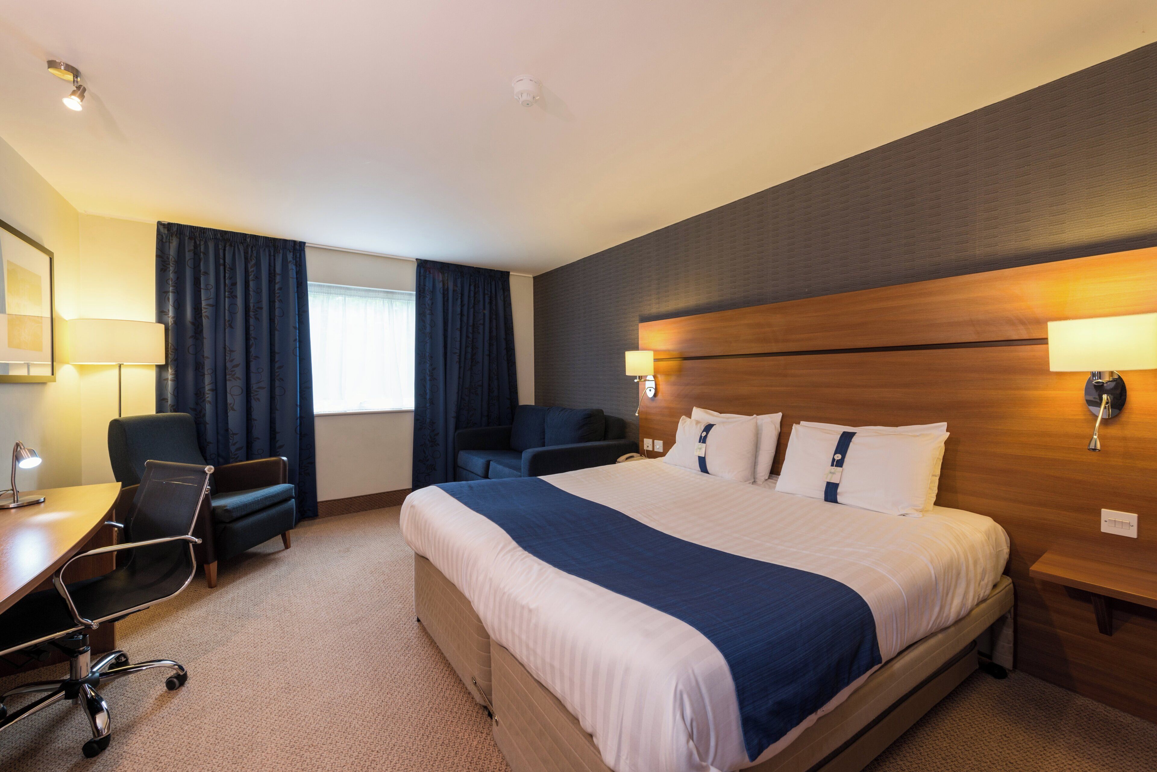 A hotel room with a large bed, two armchairs, a desk with a lamp, and a window with curtains.