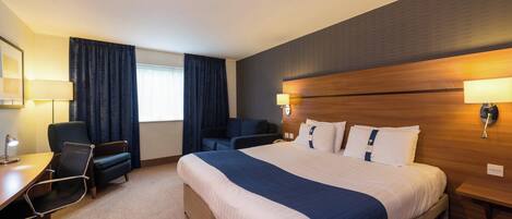 A hotel room with a large bed, two armchairs, a desk with a lamp, and a window with curtains.