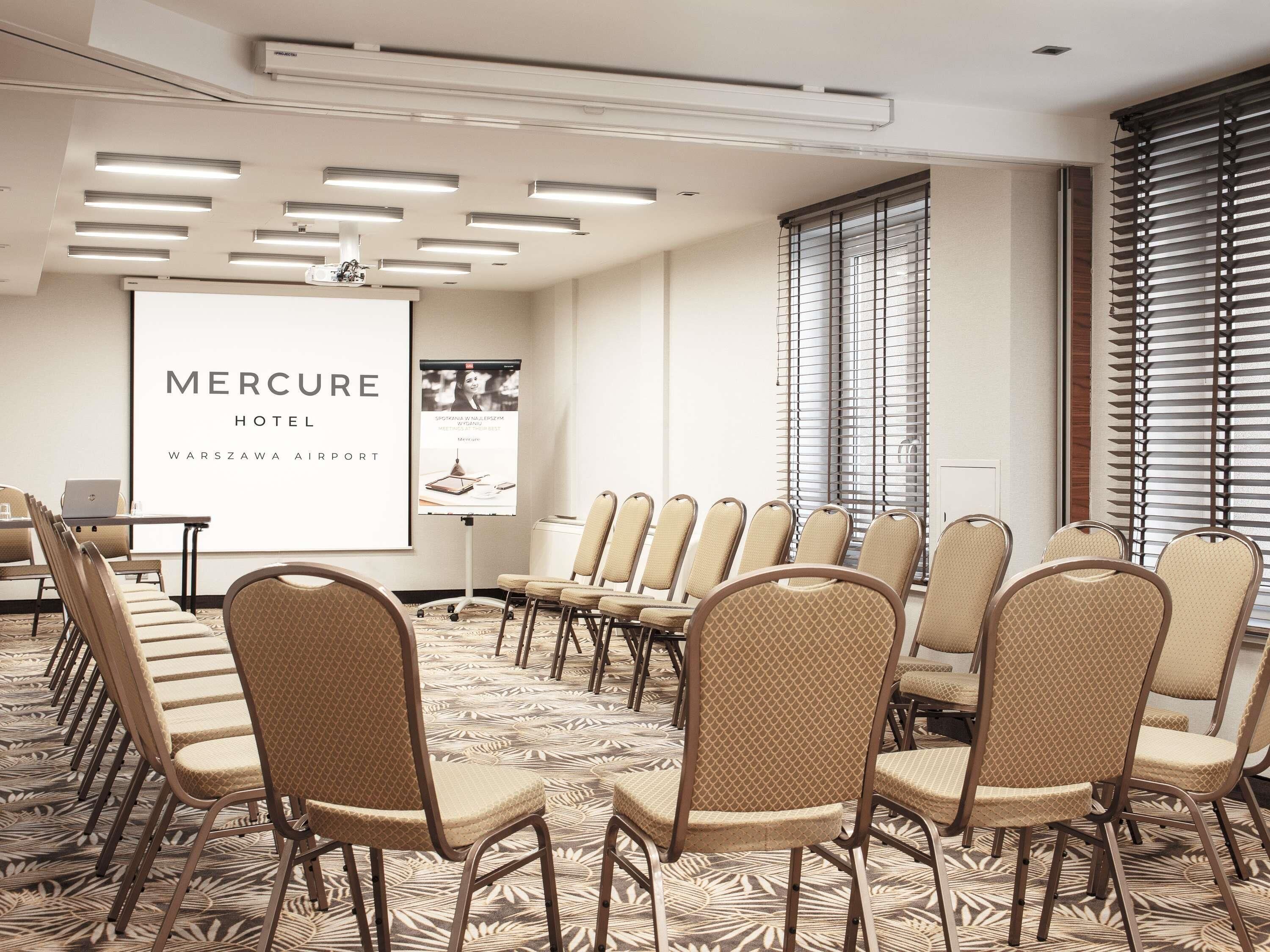 Une salle de conférence équipée d’un écran de projection affichant le nom « Mercure Hotel Warszawa Airport » et d’une affiche. La salle est dotée d’un sol recouvert de moquette, de chaises et d’une table.