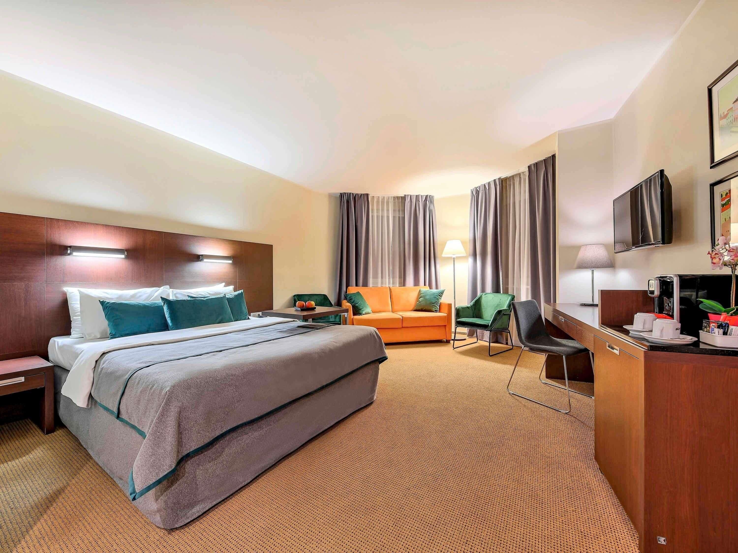Habitación de hotel con cama, escritorio, silla, sofá, cortinas, televisor y lámpara.