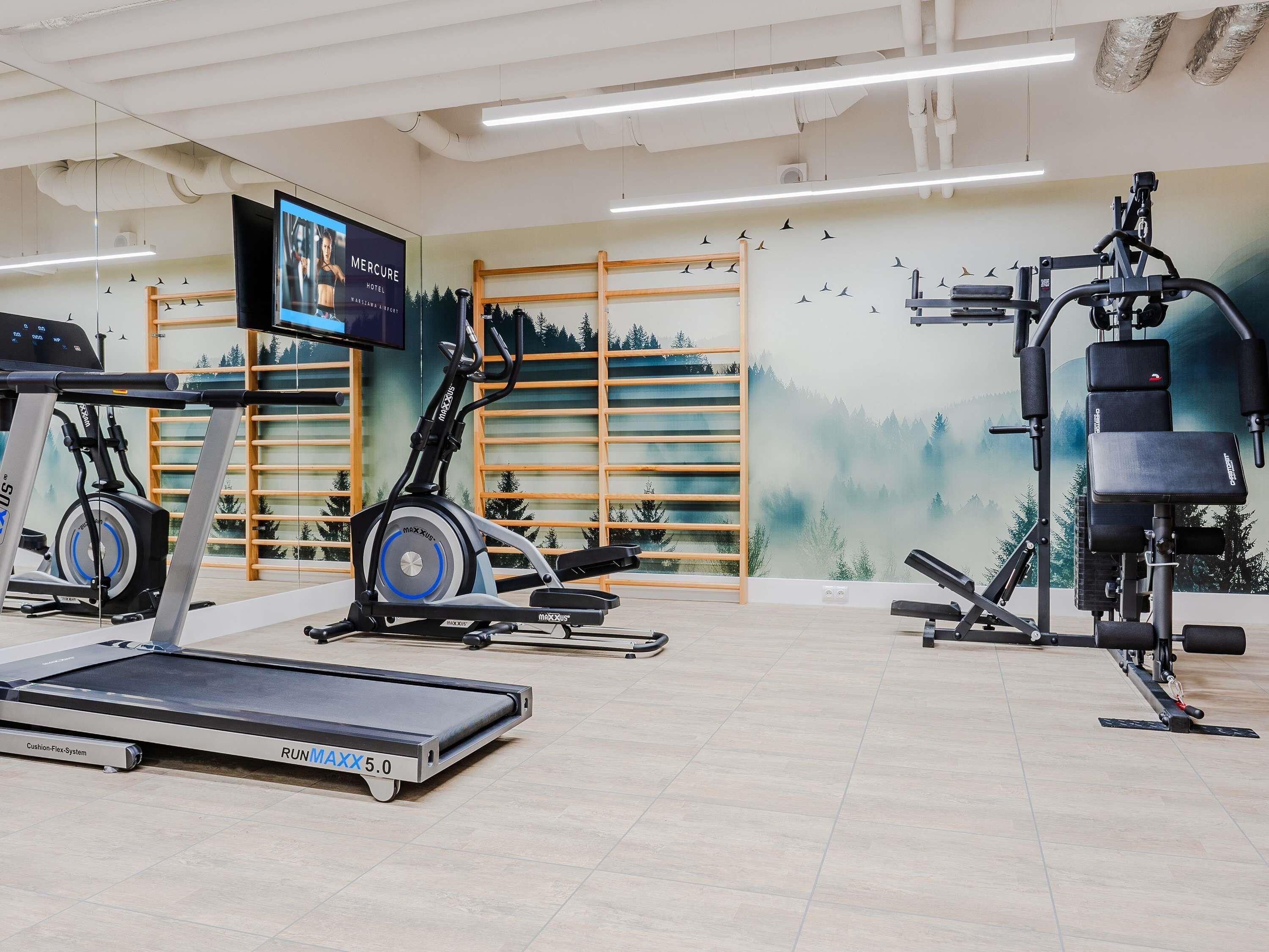 A gym with cardio equipment, a wall-mounted TV, and a mural of a forest.