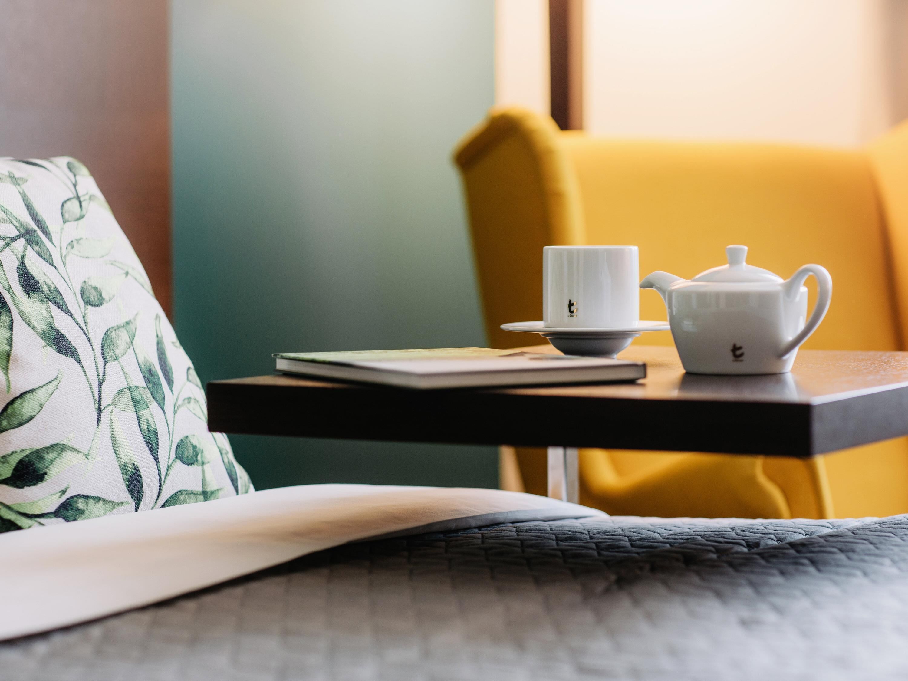 A table with a white teapot and cup, a notebook, and a green patterned pillow on a bed.