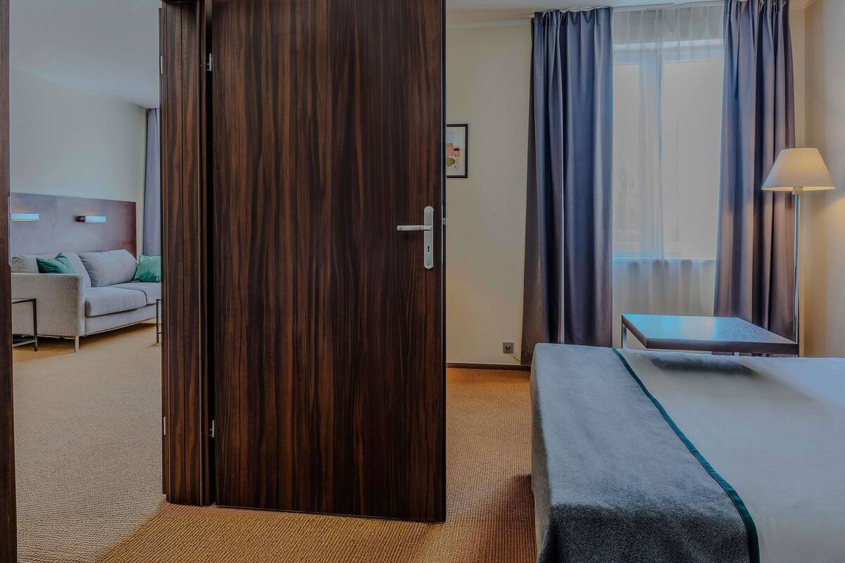 A hotel room with a wooden door, a bed with a grey bedspread, a bedside table, a lamp, a sofa, and a window with curtains.