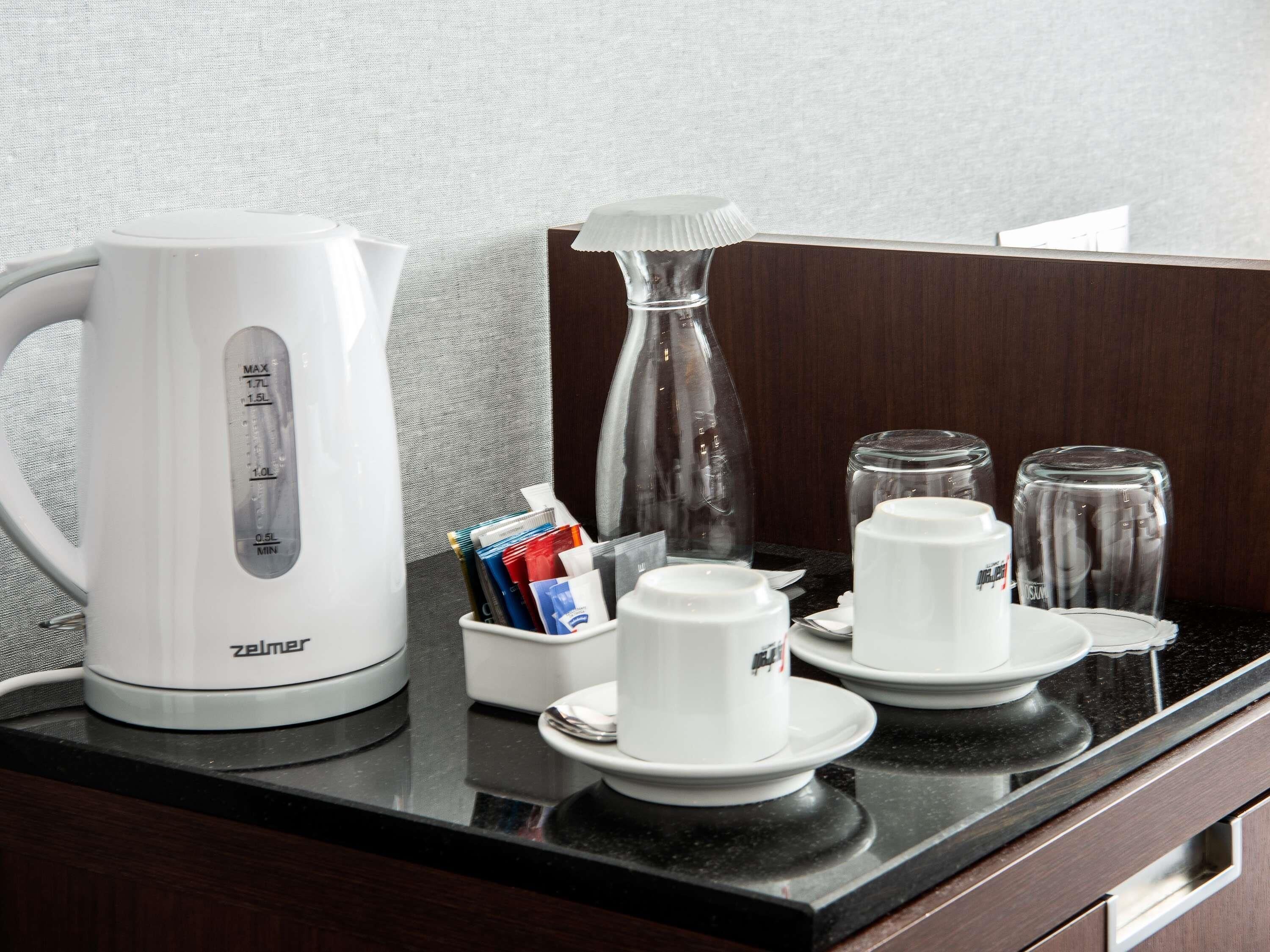 Une petite cuisine équipée d’une bouilloire blanche, d’une machine à café, d’une carafe en verre et d’une cafetière. On y trouve également deux tasses et deux verres sur le comptoir.