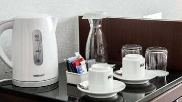 Une petite cuisine équipée d’une bouilloire blanche, d’une machine à café, d’une carafe en verre et d’une cafetière. On y trouve également deux tasses et deux verres sur le comptoir.