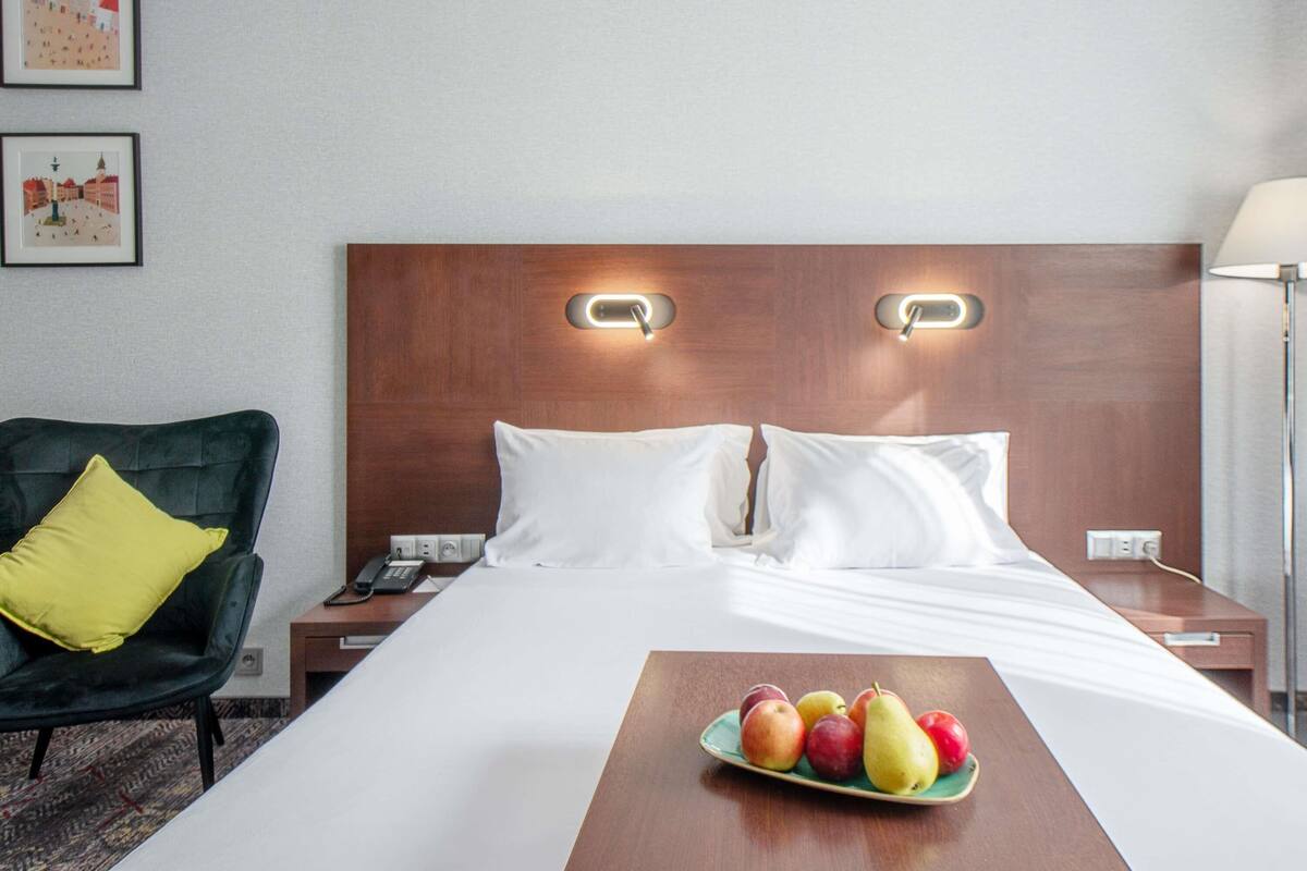 A hotel room with a bed, bedside tables, a chair, and a fruit bowl on the nightstand.