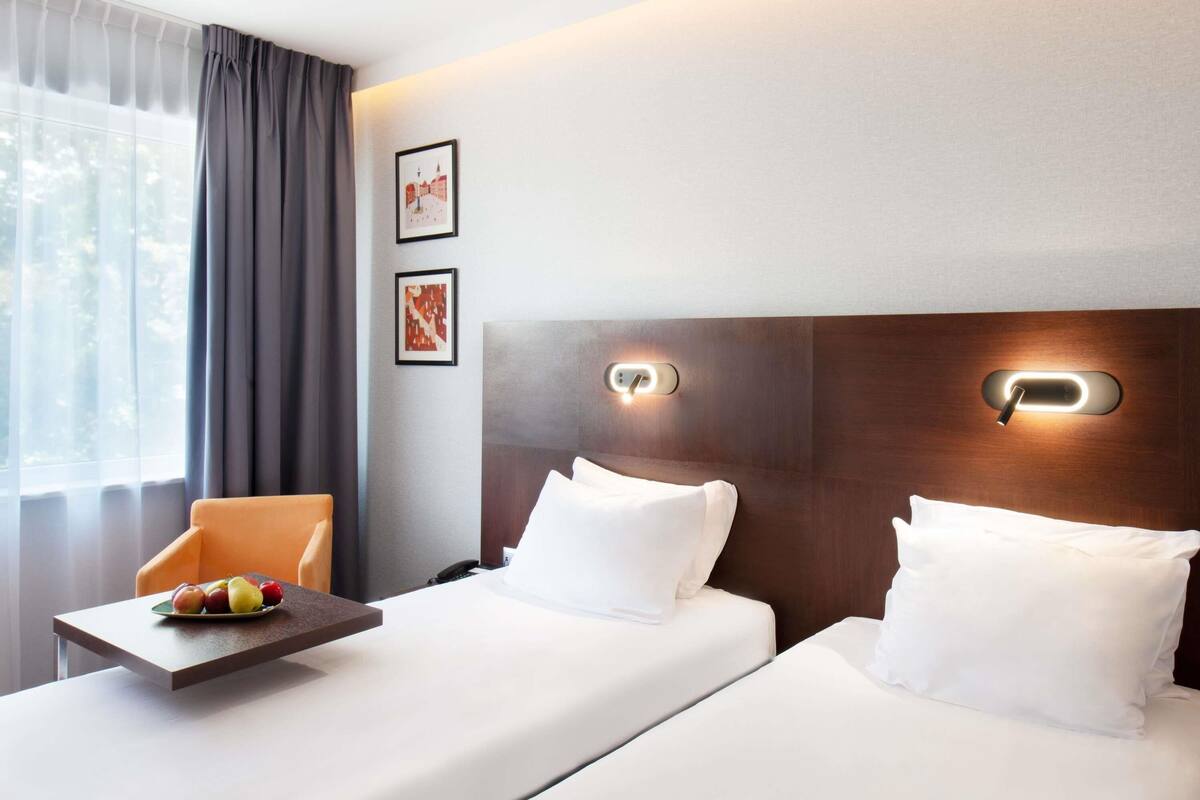 A hotel room with two beds, a small table with fruit, a chair, and two framed pictures on the wall.