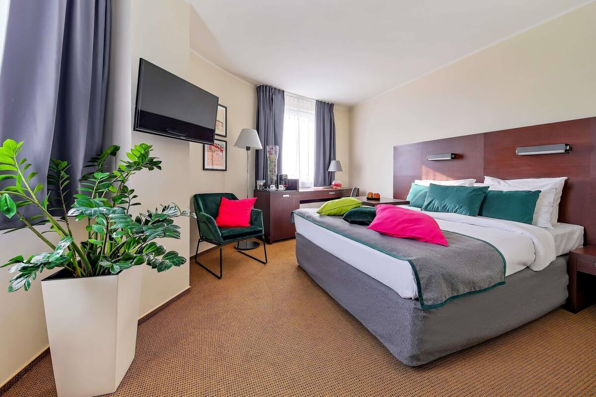 A hotel room with a bed, a desk, a chair, a TV, and a potted plant.