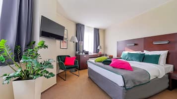 Et hotelværelse med en seng, et skrivebord, en stol, et fjernsyn og en potteplante.