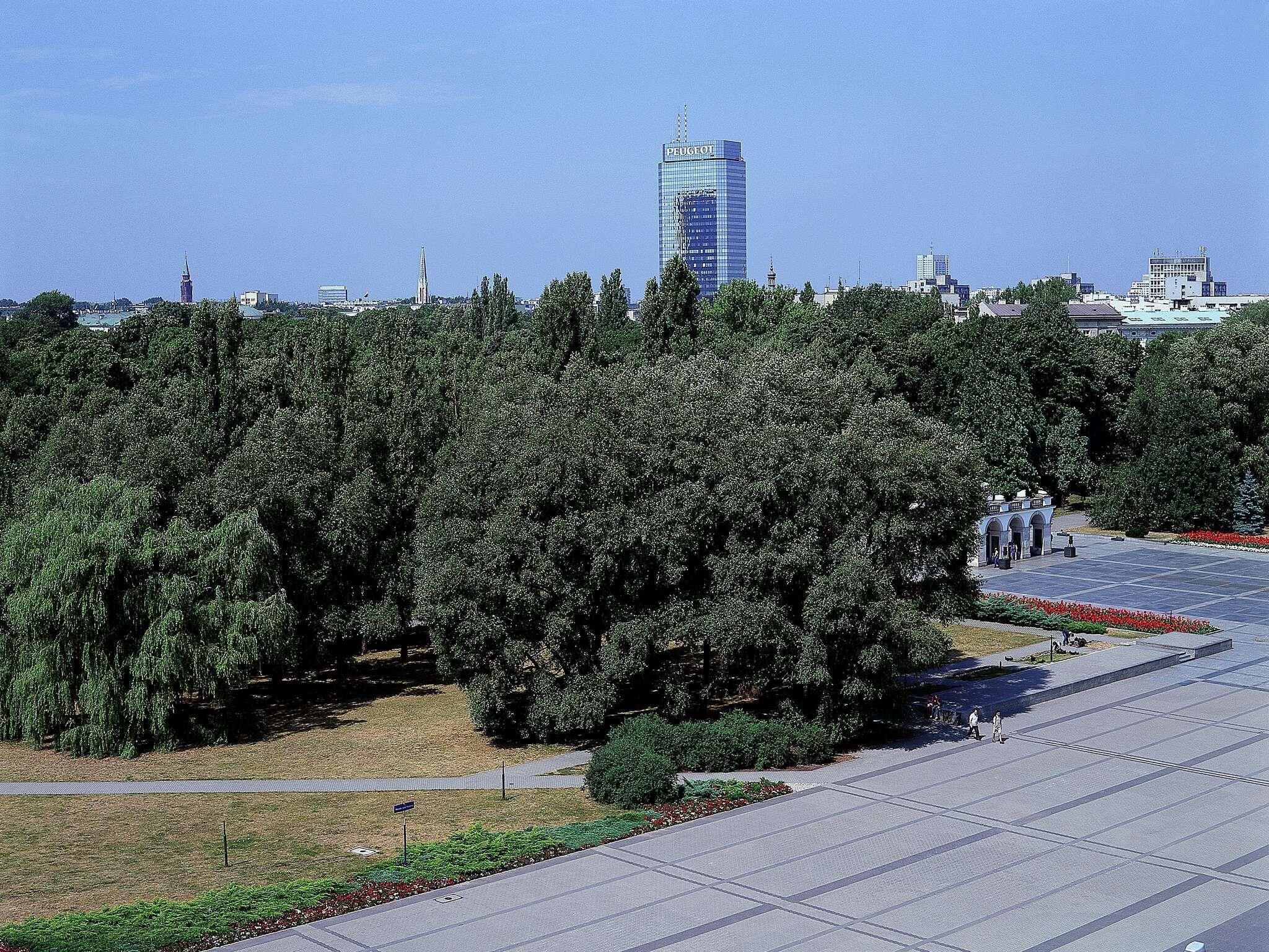 Une vaste place ouverte, avec un grand immeuble en arrière-plan, entourée d’arbres denses.