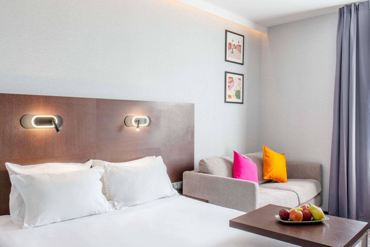 A hotel room with a bed, a sofa, a small table with fruit, and wall-mounted light fixtures.