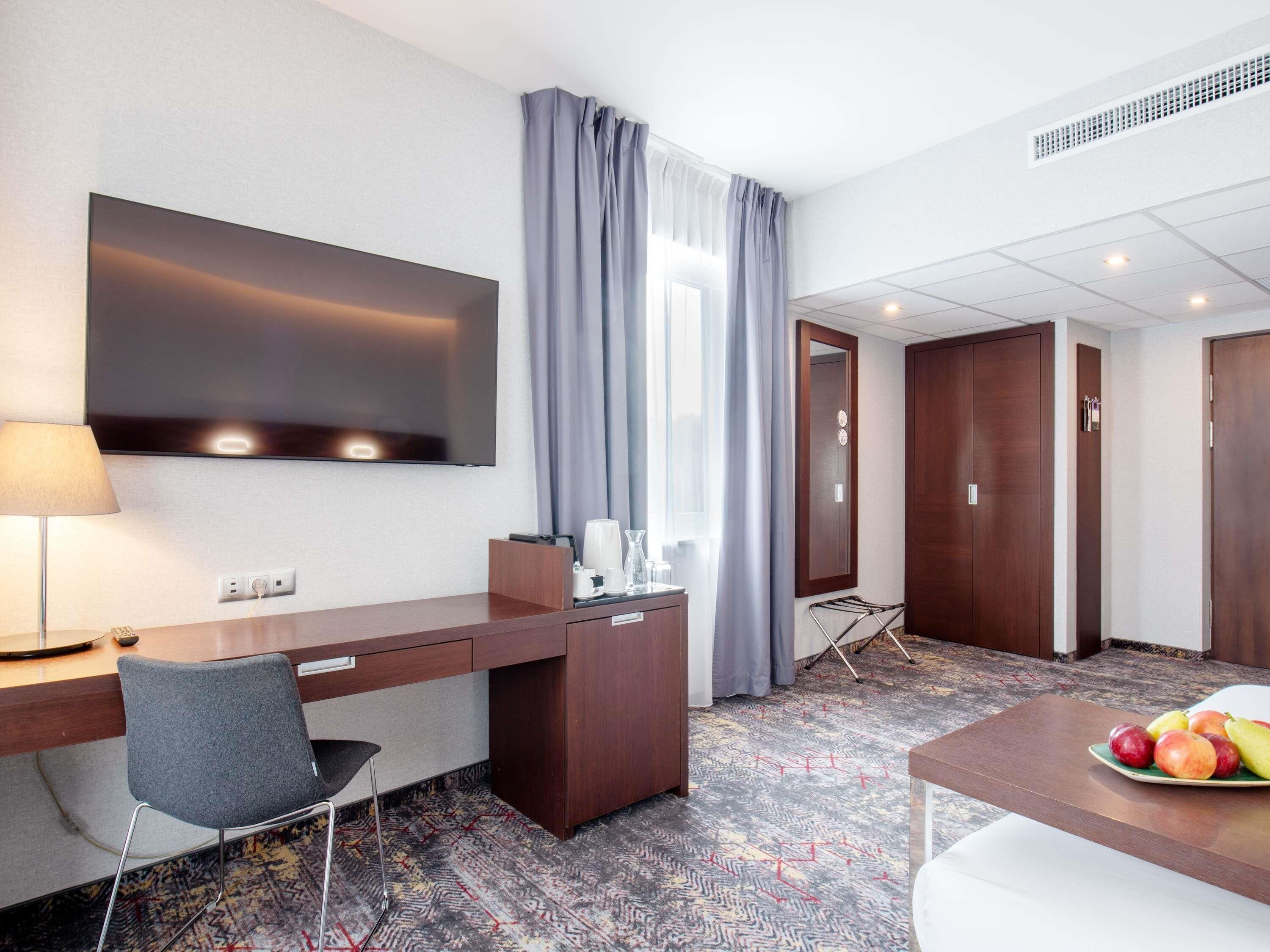 A hotel room with a desk, chair, TV, and a dining area with fruit.