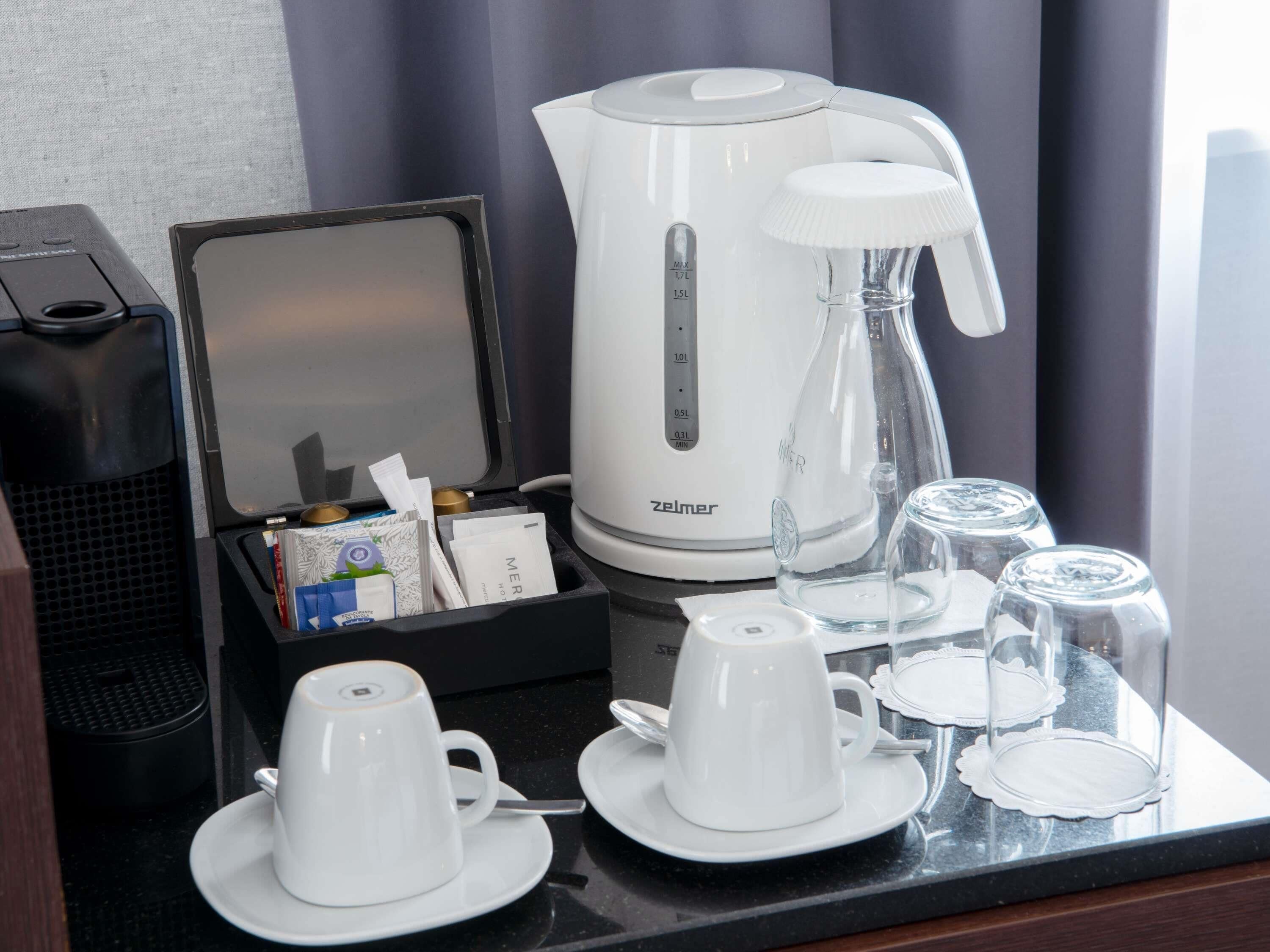 A hotel room with a coffee maker, tea kettle, cups, and glasses on a table.