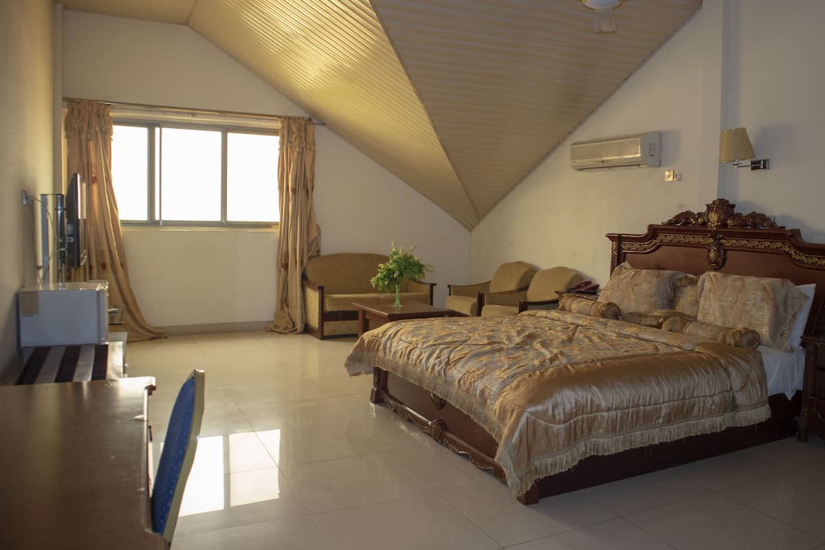 Une chambre spacieuse avec un grand lit, un bureau et une chaise. Elle est dotée d’une fenêtre avec des rideaux, d’un ventilateur de plafond et d’un climatiseur fixé au mur.