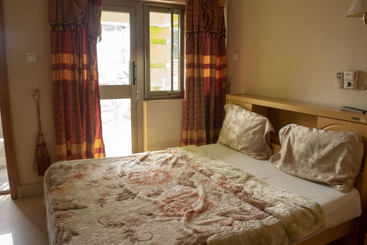 Un lit avec une couvre-lit à motifs, une tête de lit en bois et deux oreillers. Il y a une fenêtre avec des rideaux, une table de chevet avec une lampe et un téléphone fixé au mur.
