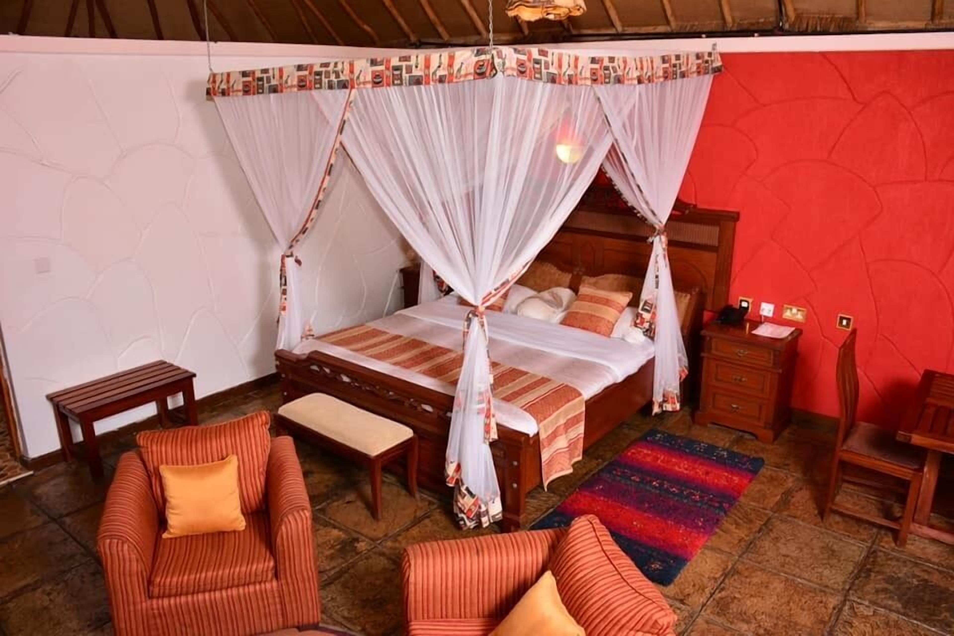 Quarto com cama sob dossel, um banco de madeira, uma mesinha, uma cadeira e um tapete colorido.