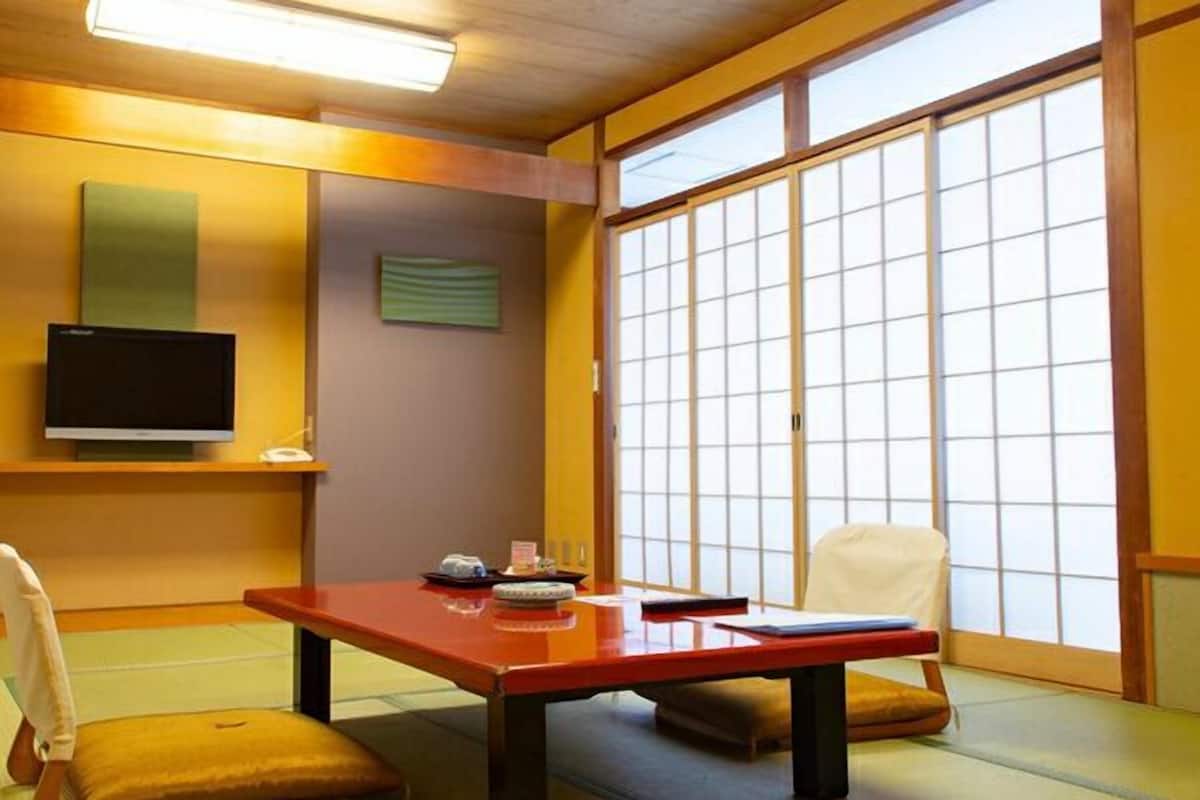 Una habitación japonesa tradicional con suelo de tatami, una mesa baja, un televisor en un estante y una puerta corredera con diseño de cuadrícula.