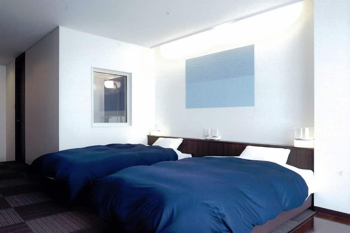 Habitación de hotel con dos camas, un ventanal grande, una lámpara de pared y una mesita de noche con lámpara.