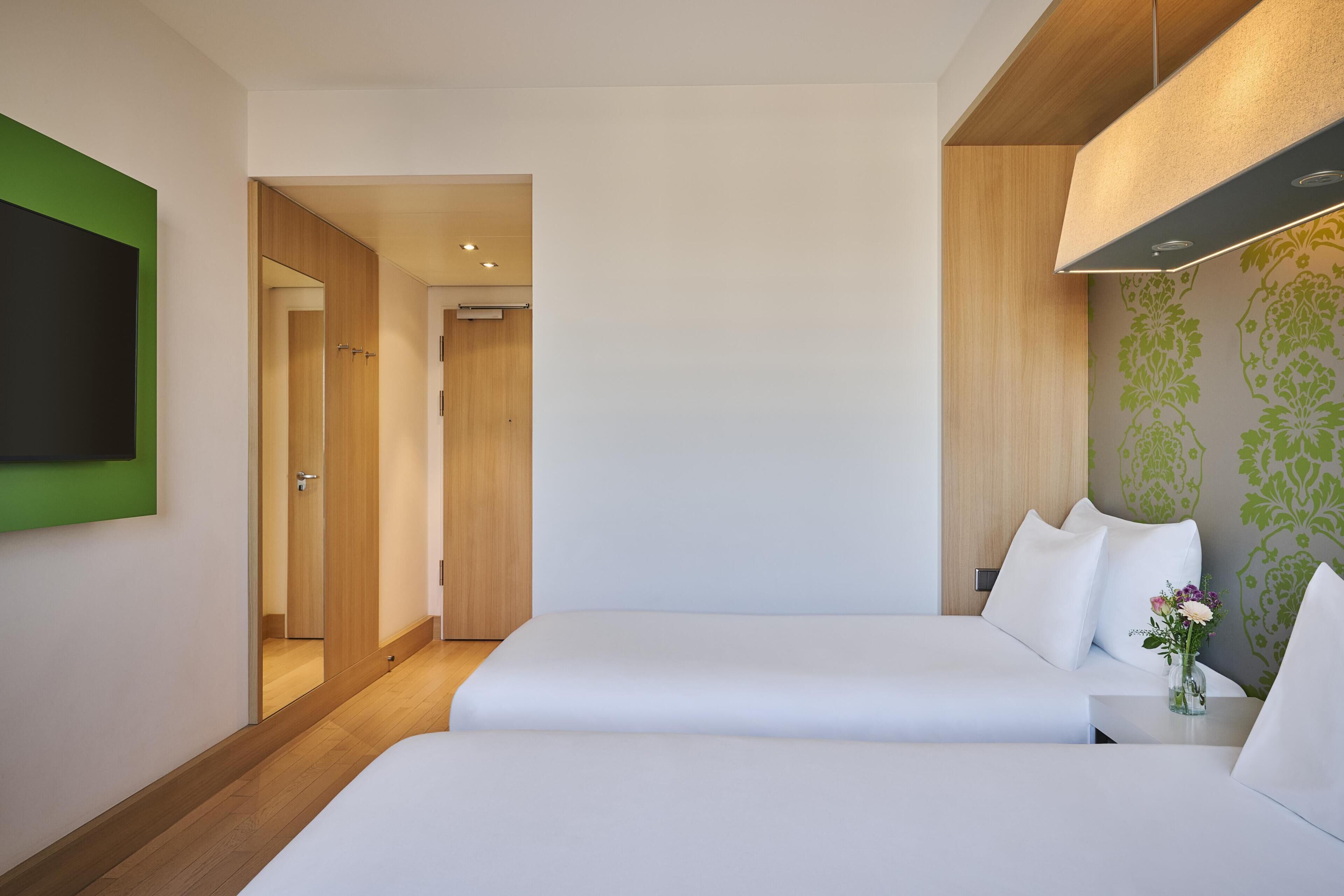 A hotel room with two beds, a flat-screen TV mounted on the wall, a wooden door, and a green patterned wall.