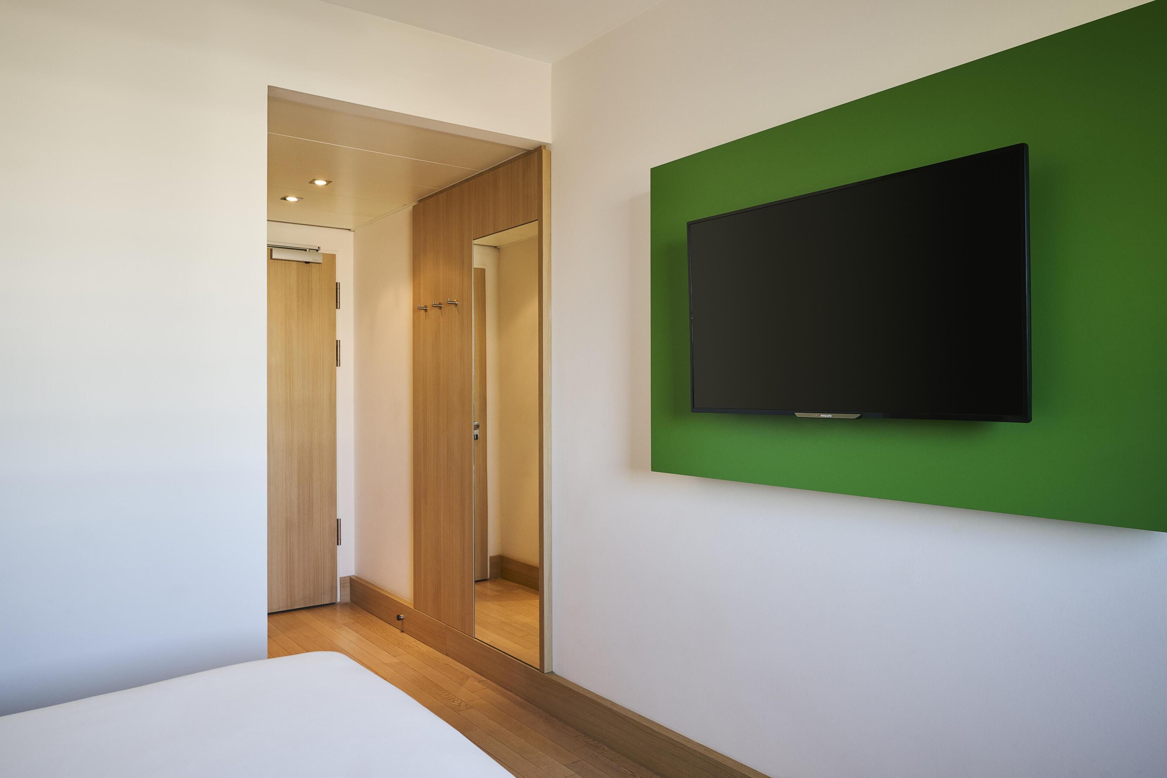 A modern hotel room with a bed, a green accent wall, a mounted TV, and a wooden door.