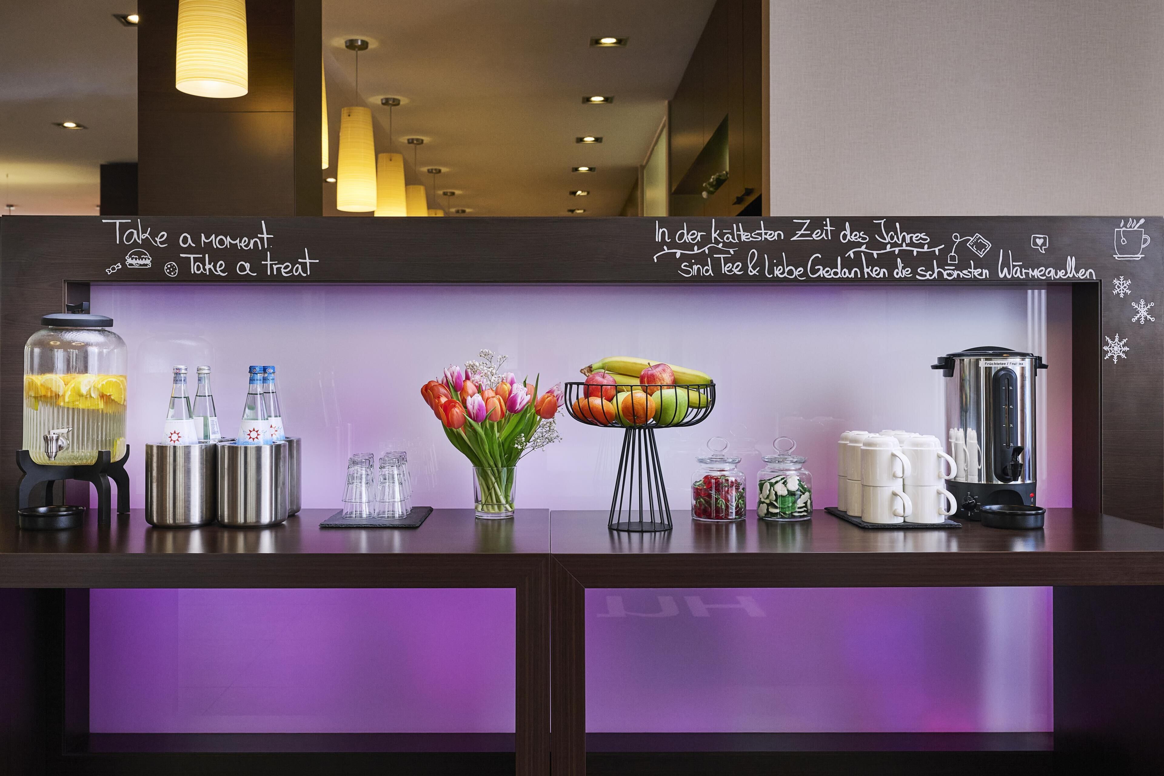 A hotel reception desk with a chalkboard displaying messages, a vase of tulips, a fruit bowl, and various amenities like a coffee maker and jars.