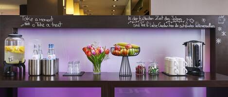 A hotel reception desk with a chalkboard displaying messages, a vase of tulips, a fruit bowl, and various amenities like a coffee maker and jars.