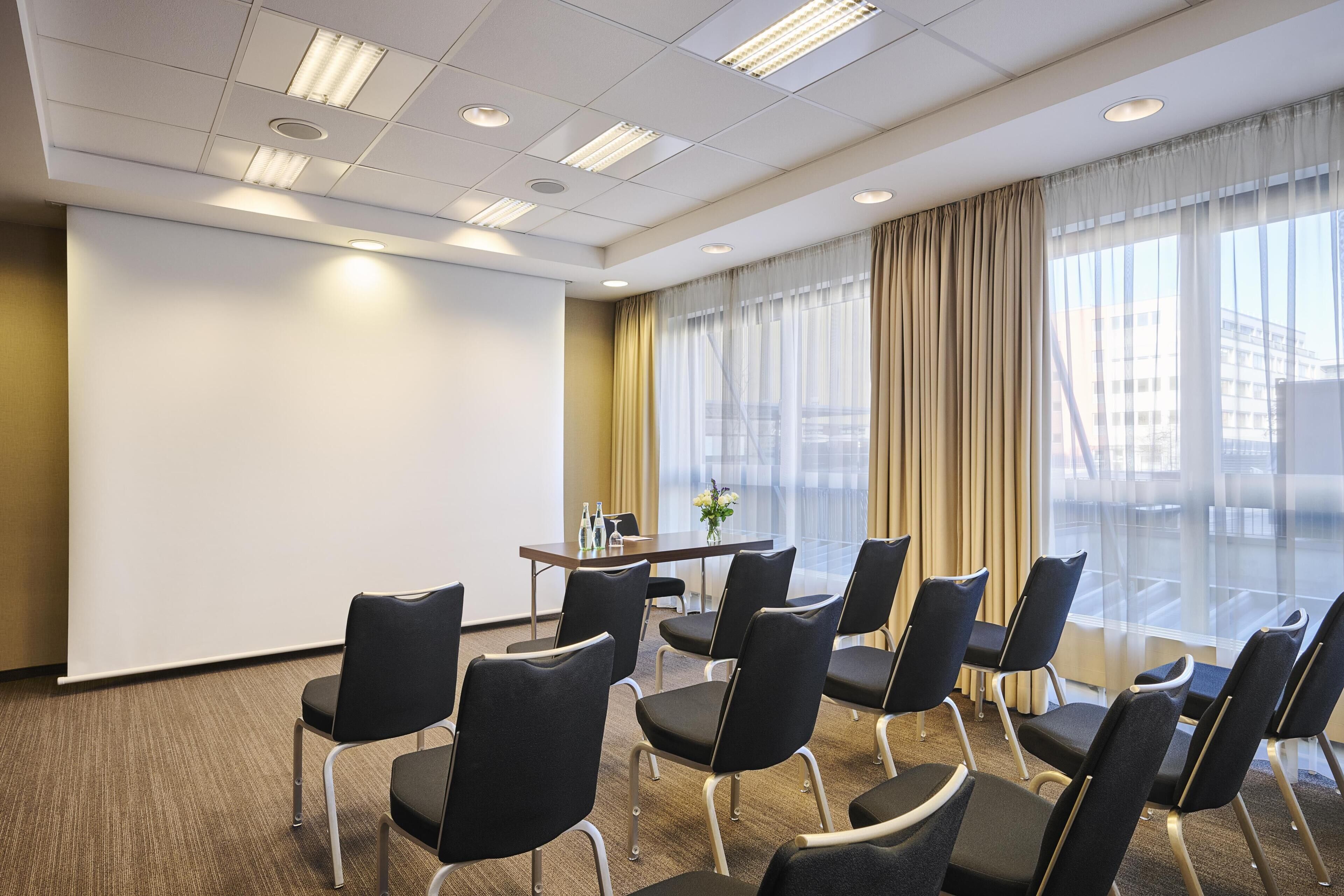 A conference room with a table, chairs, a projector screen, and a large window with curtains.