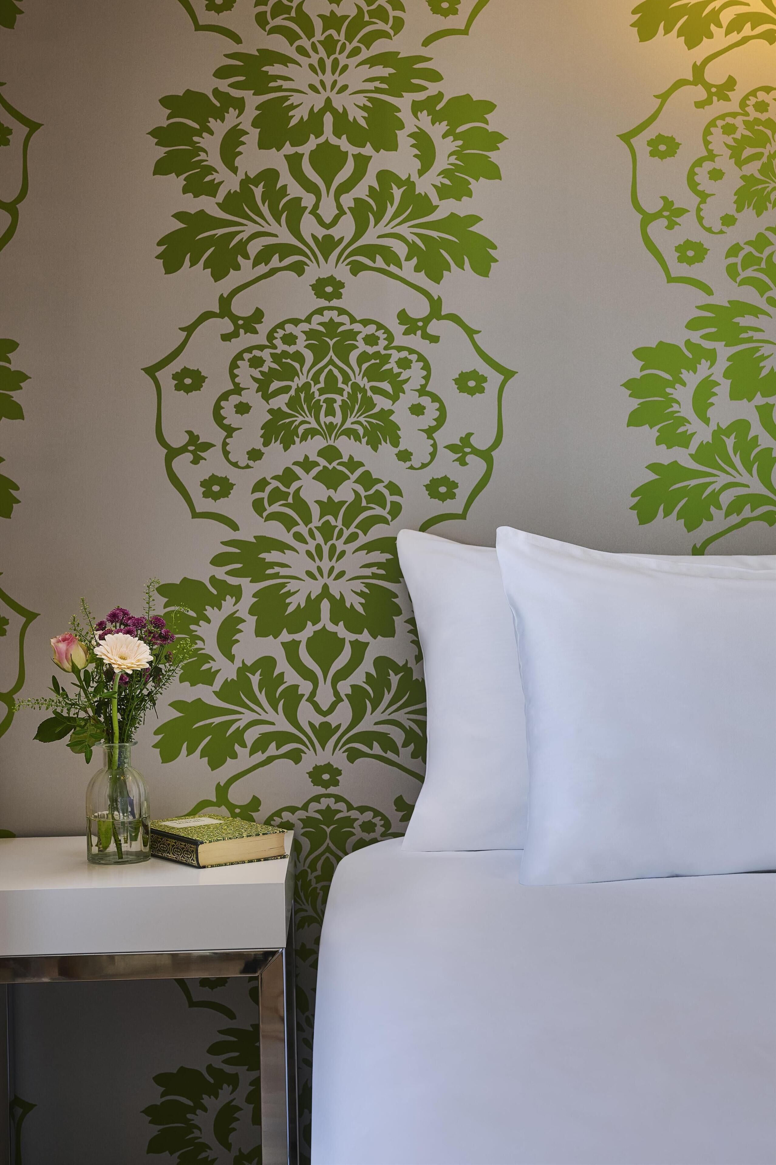 Una cama con ropa de cama blanca, una mesita de noche con un florero y un papel tapiz estampado.
