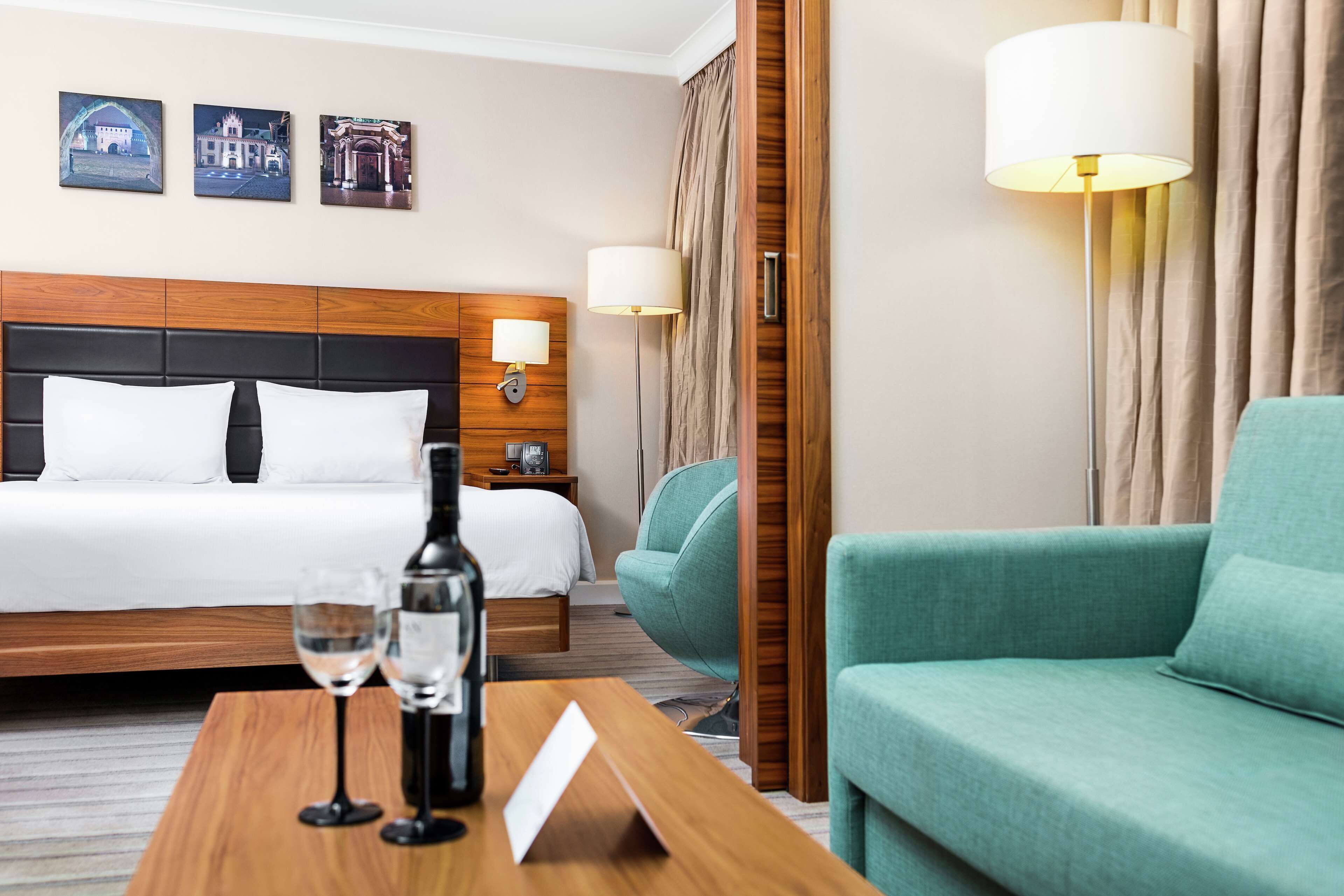 A hotel room with a bed, a wooden headboard, a green armchair, a wooden table with a bottle and glasses, and a wall-mounted lamp.
