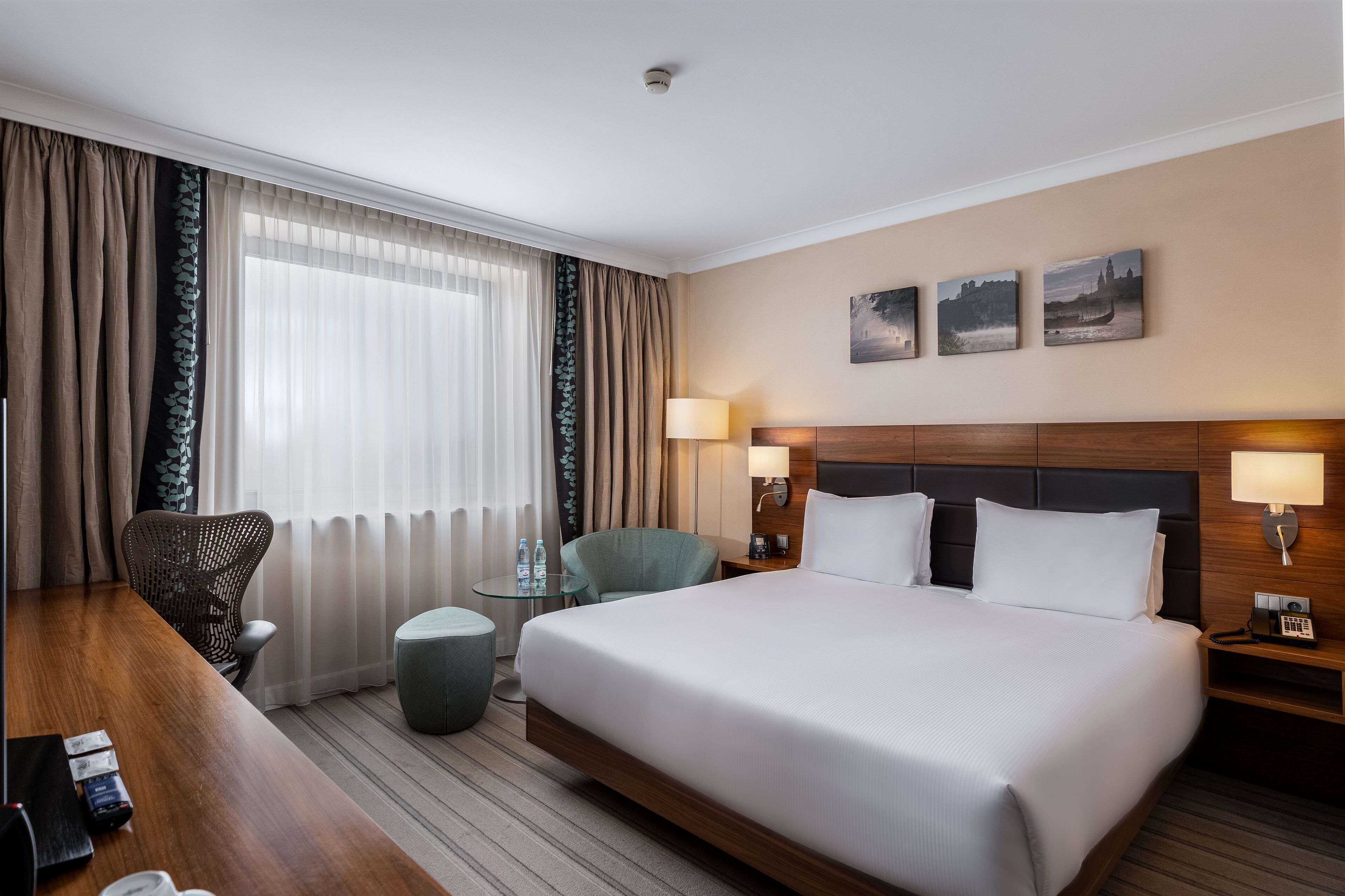 A hotel room with a large bed, two bedside tables with lamps, a desk with a chair, a round ottoman, and a view of a cityscape through the window.