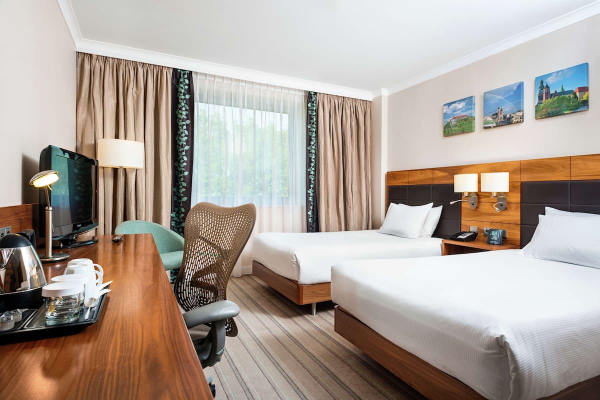 A hotel room with two beds, a desk with a lamp, a chair, a TV, and a tray with cups and glasses.